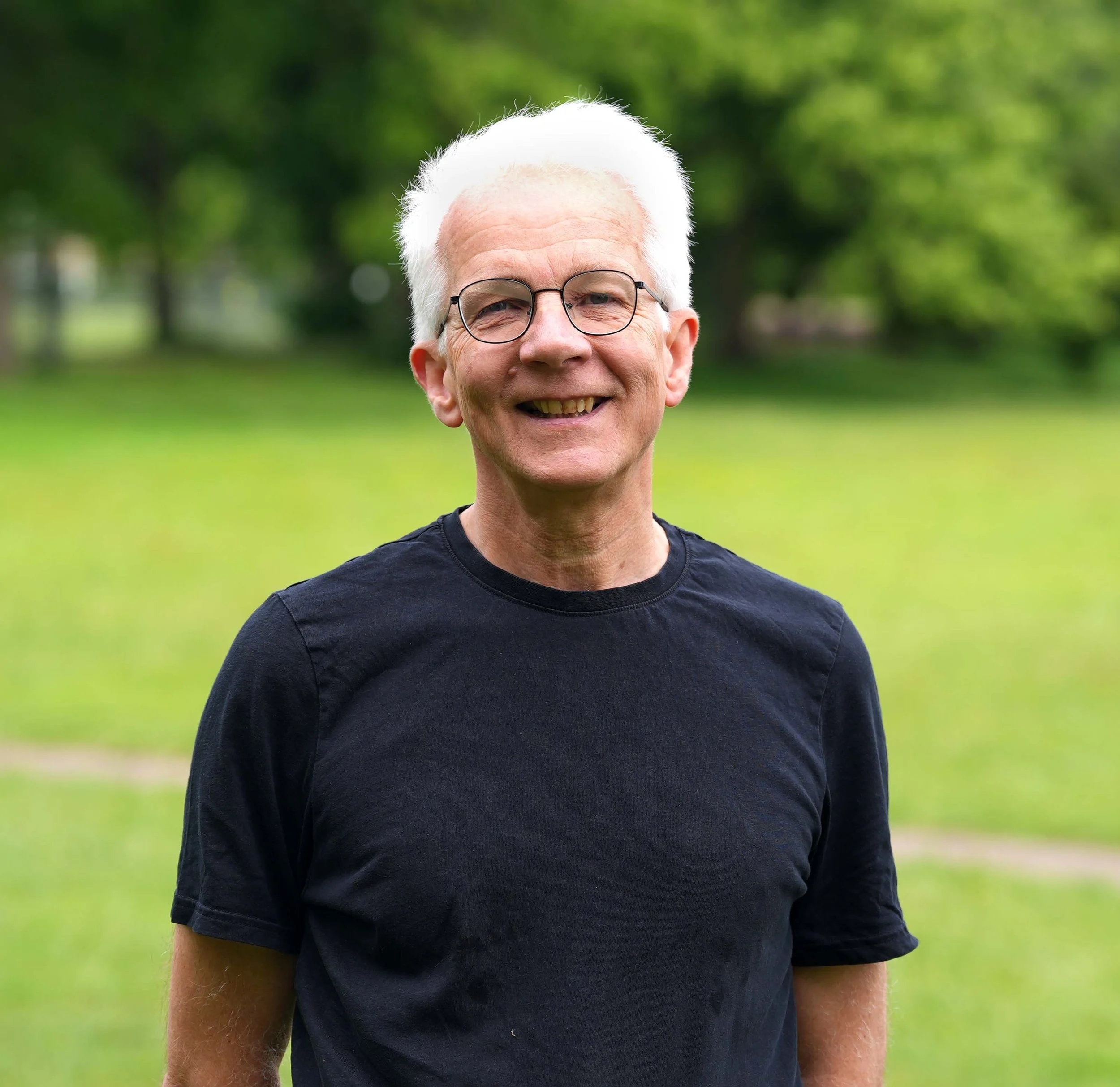Close-up of smiling elderly man with white hair and glasses outdoors in a park, green trees in background.