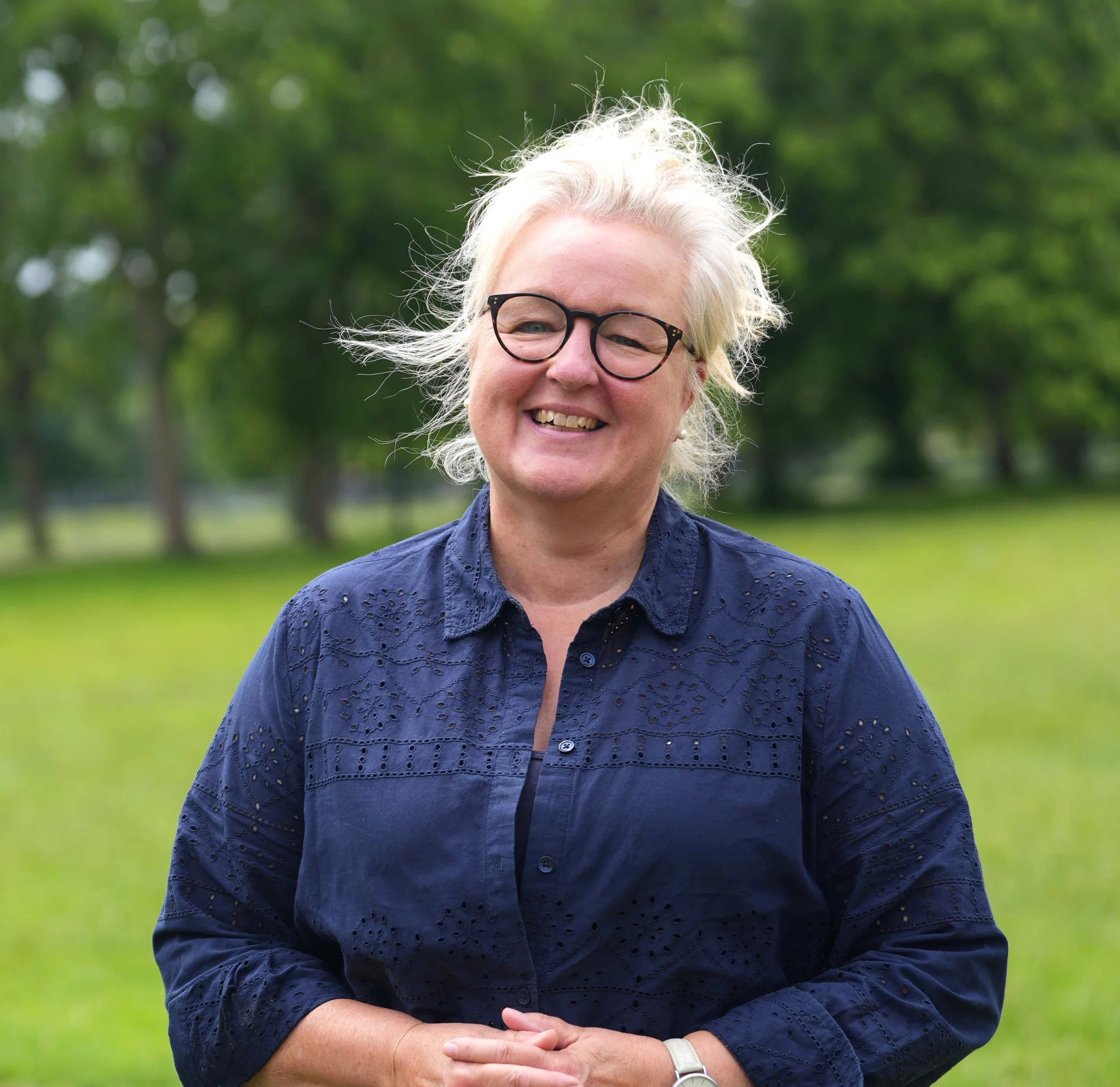 Close-up of a smiling woman with short, curly, platinum blonde hair and glasses, wearing a dark blue embroidered blouse, standing outdoors with out-of-focus greenery in the background.
