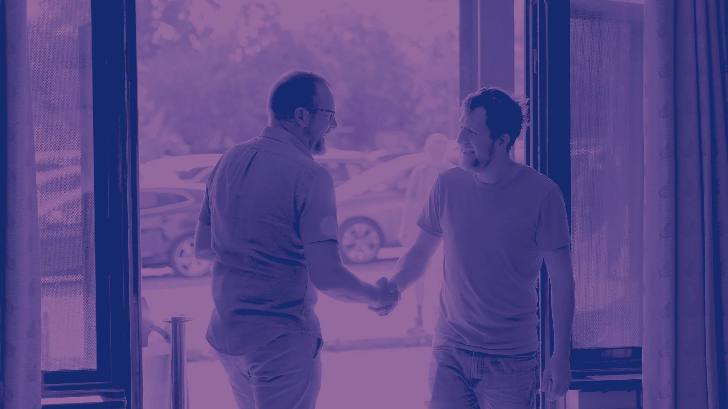 Two men shaking hands and smiling at each other at the entrance of a building, with cars visible outside.