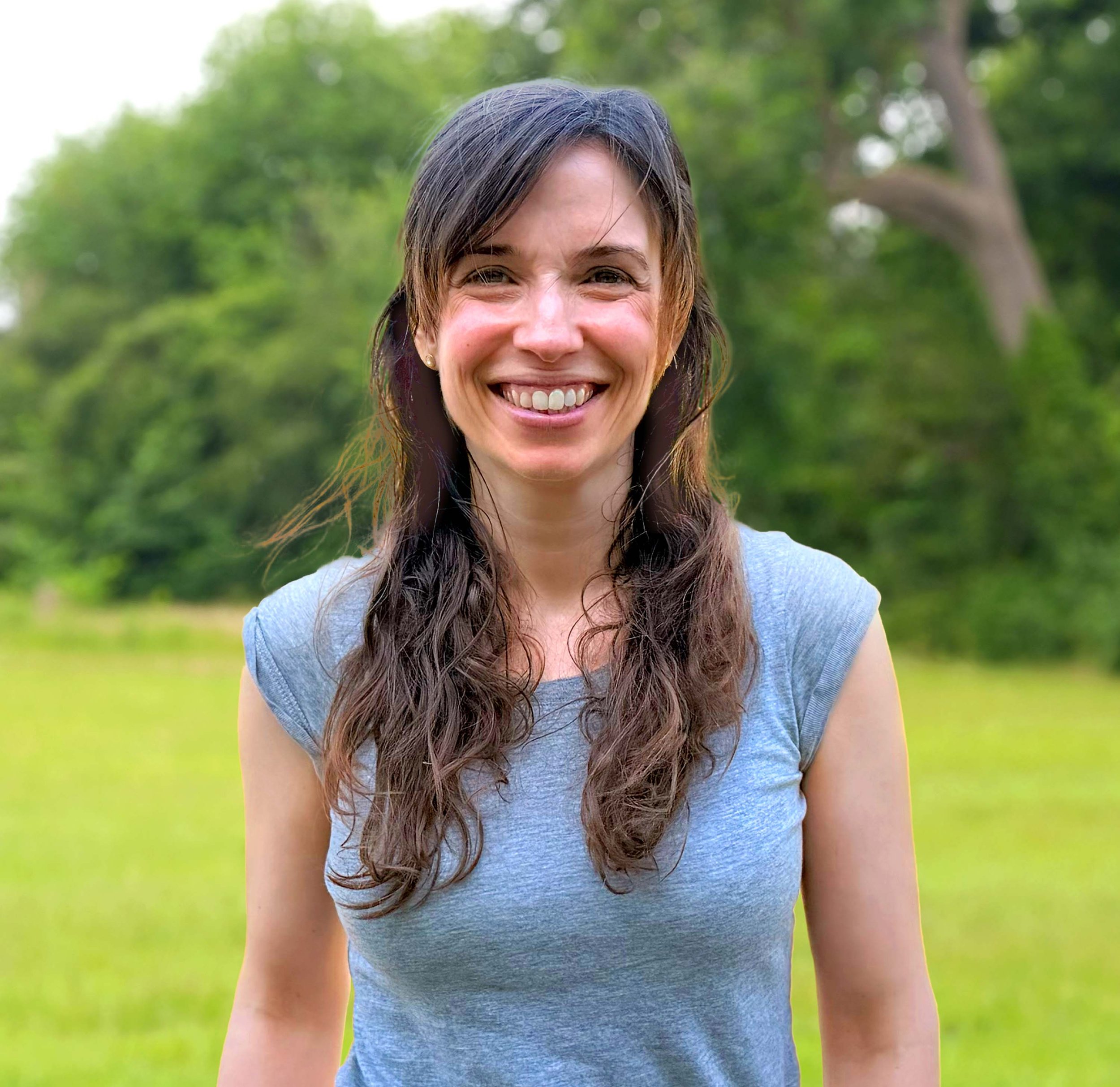 Close-up of a smiling woman with long, wavy brown hair, wearing a light blue shirt, outdoors with blurred green trees in the background.