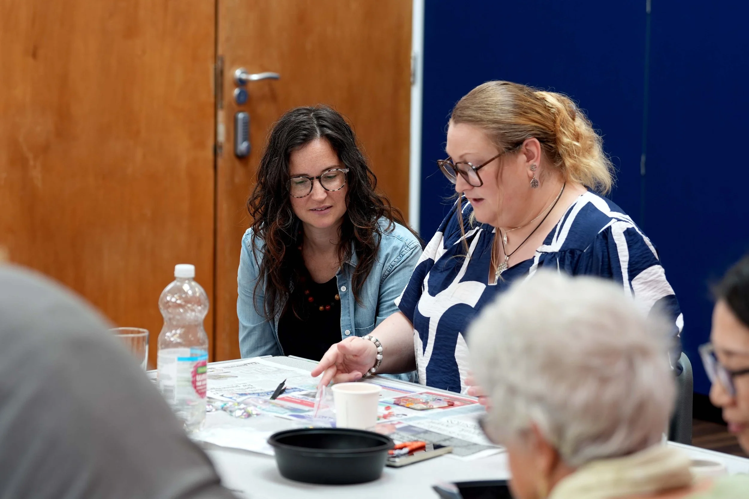 Two women sitting at a table, engaged in conversation, with various papers and drinks, including water bottles and a cup, in front of them.