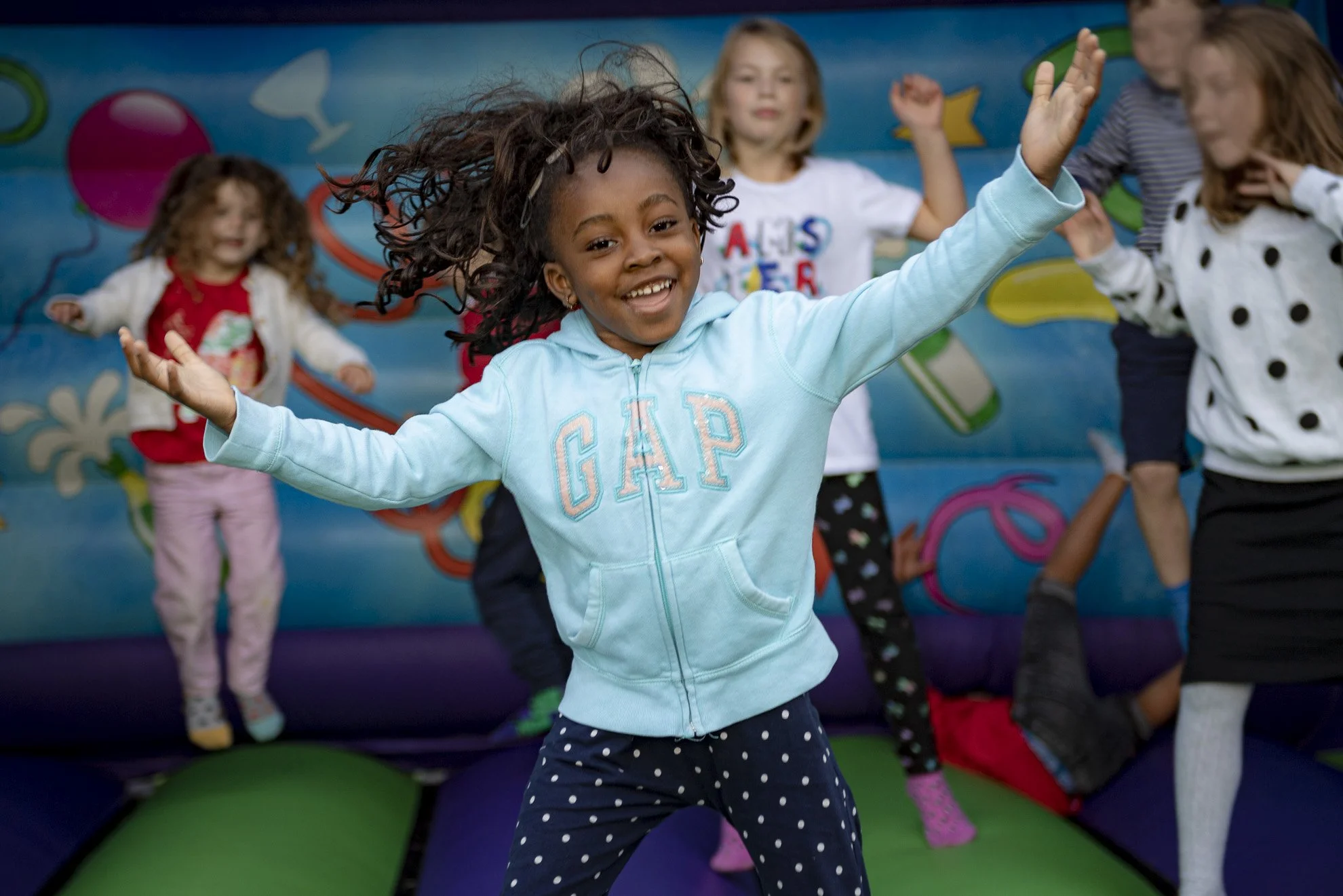 Young girl with curly hair dancing and smiling in a colorful indoor play area with other children around her.