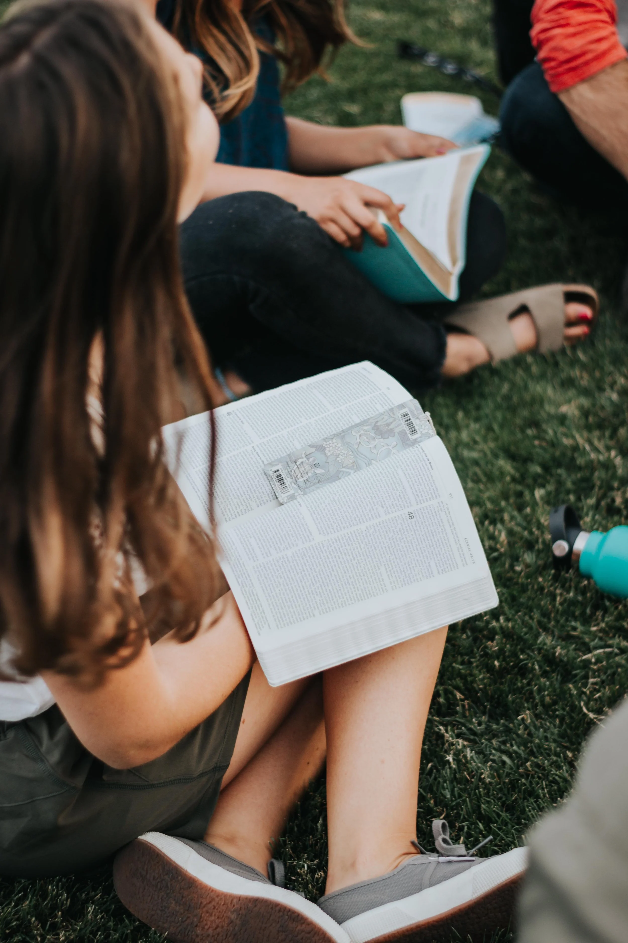 A person with long brown hair reading a Bible while sitting on grass outdoors, with other people nearby also reading.