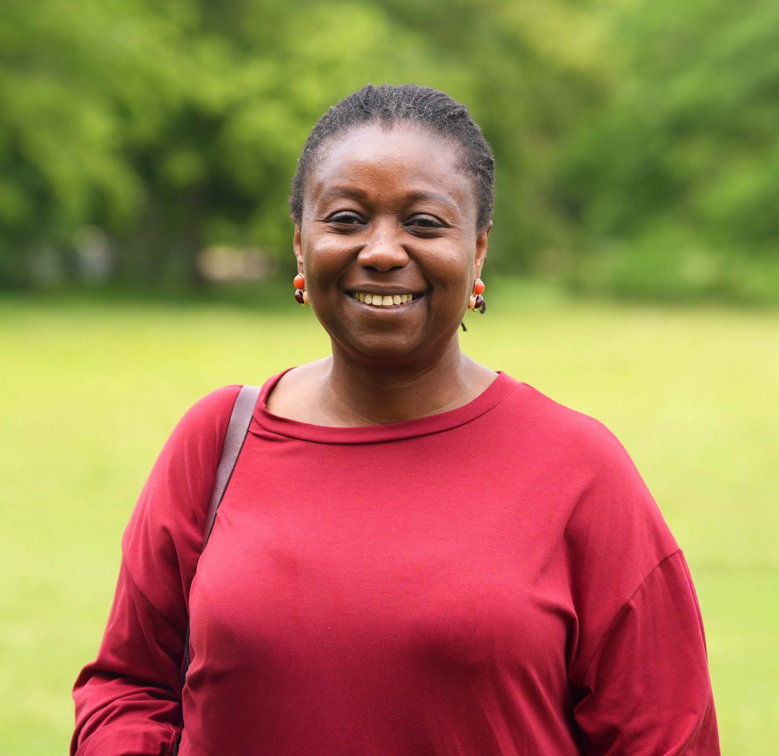 Close-up of a smiling African American woman with short curly hair, wearing a red top and earrings, outdoors with blurred green background.