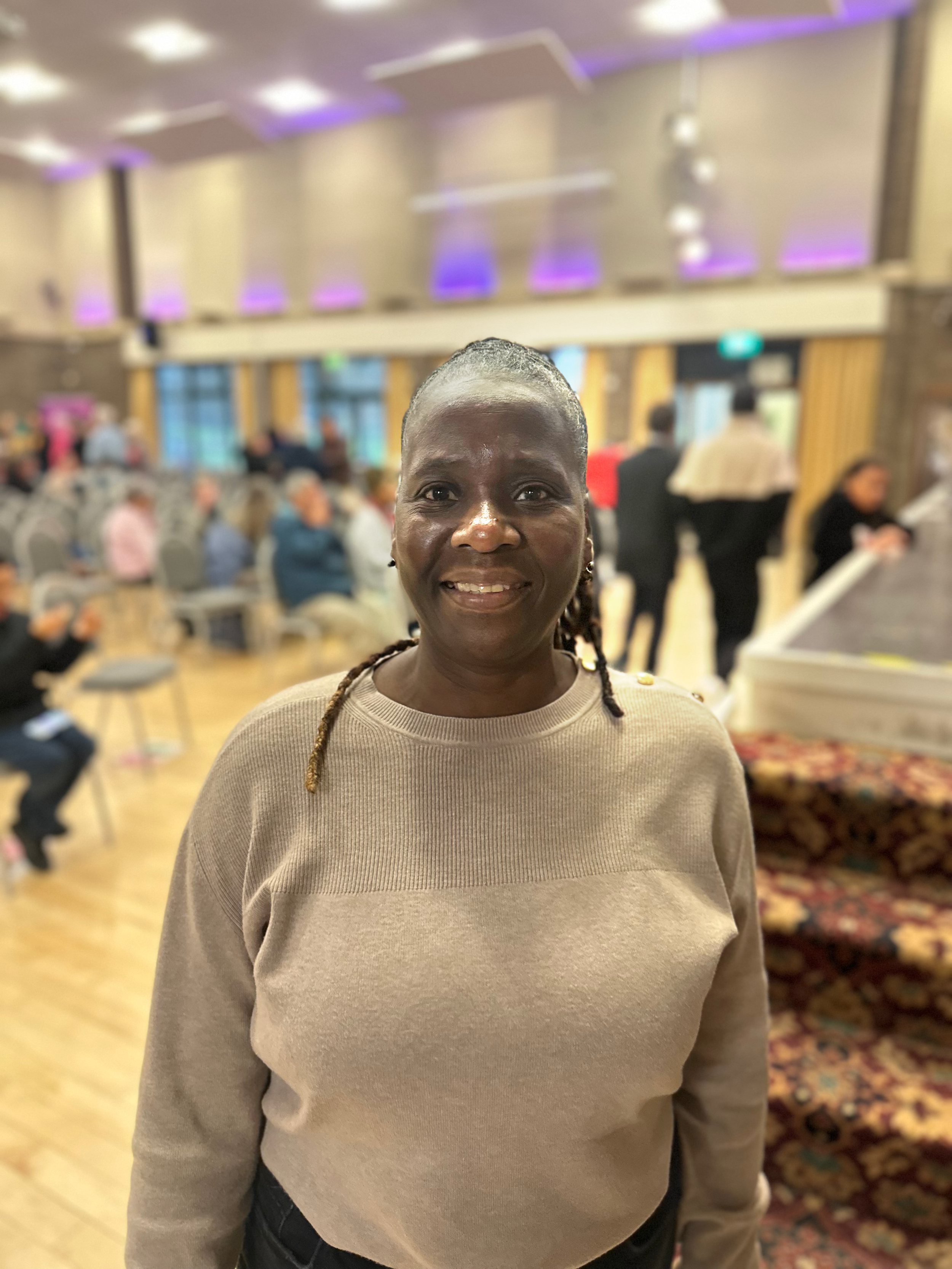 A woman with braided hair smiling at the camera, standing in a large indoor event space with many people, chairs, and tables in the background.