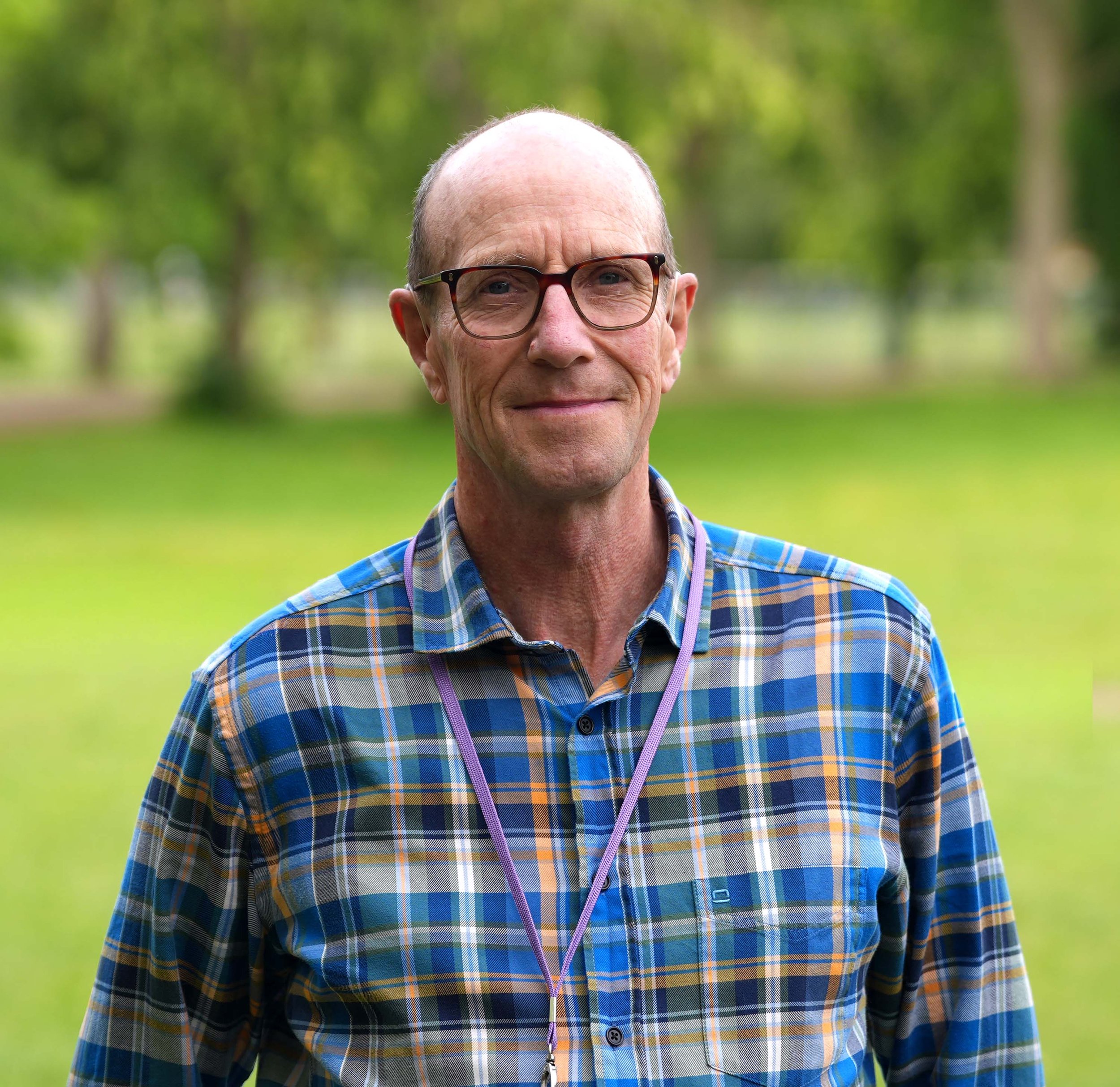 Close-up of a middle-aged man with glasses and a plaid shirt smiling outdoors with green trees in the background.