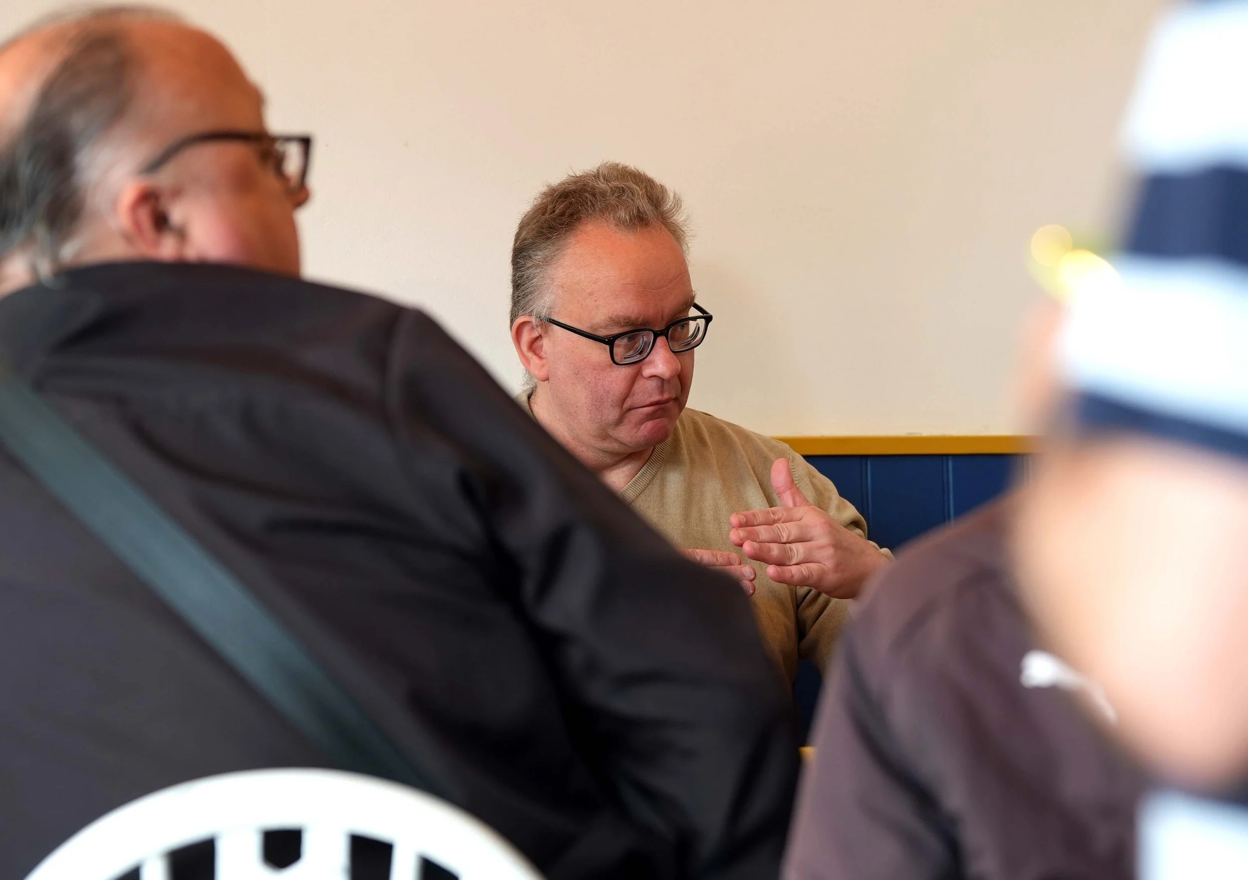 Two men are sitting indoors, one wearing glasses and a beige sweater with a serious expression, and the other in a leather jacket with glasses, partially visible in the foreground.