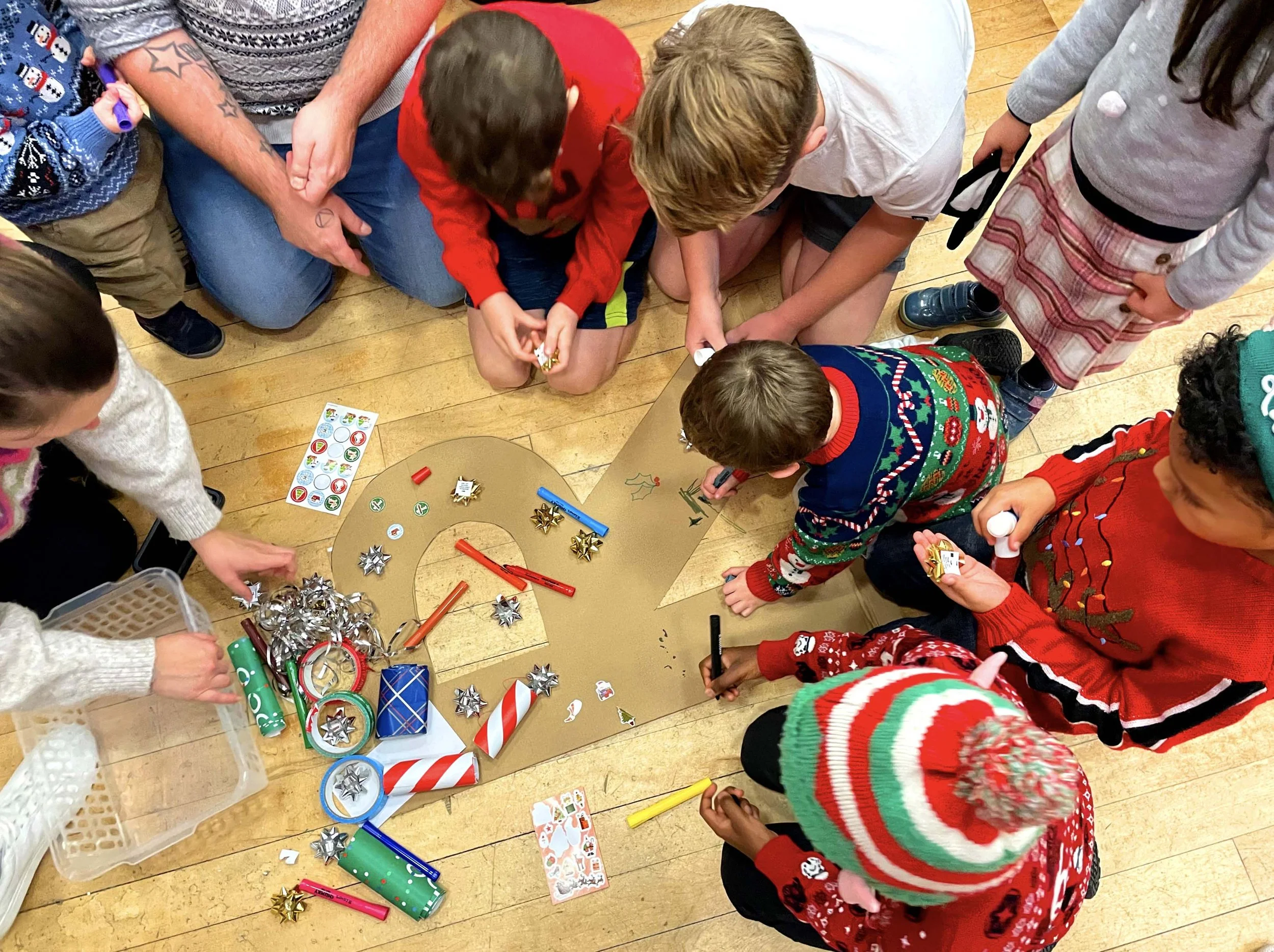 Children and adults gathered around a table, decorating with Christmas-themed items, such as bows, stickers, and wrapping paper, during a festive celebration.