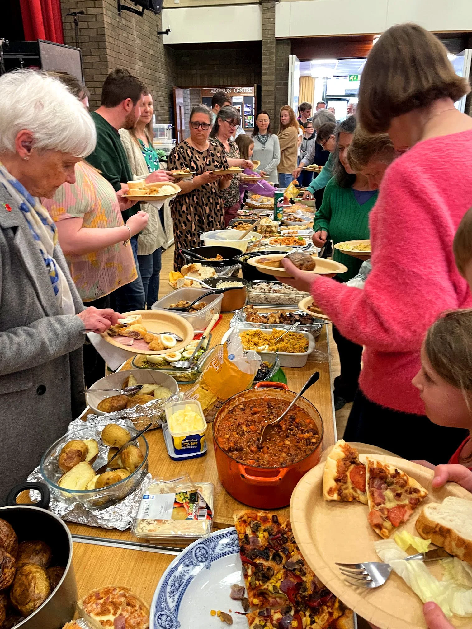 People standing in line at a buffet table with various food dishes such as pizza, chili, salads, and baked potatoes in a hall or dining area. Big Sunday Lunch at Grace