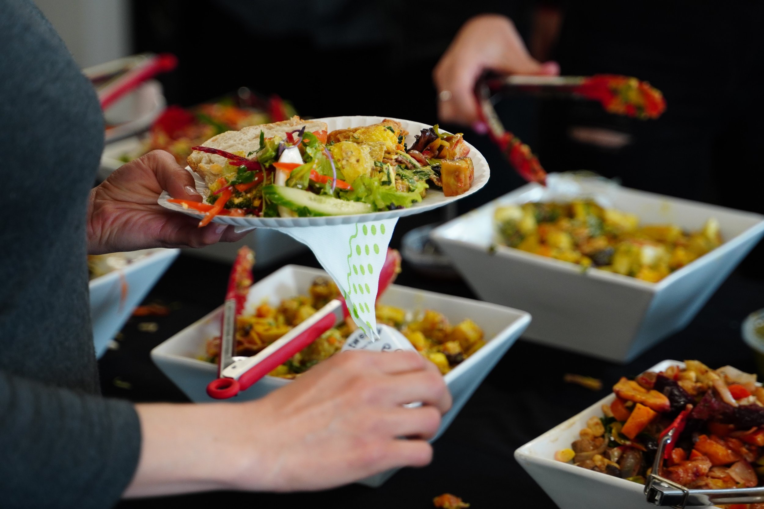 Person holding a paper plate filled with salad and food at a buffet with various dishes in the background.