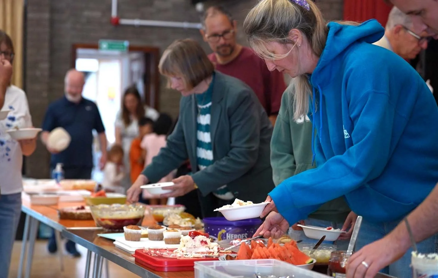 People standing in a line serving themselves food on a buffet table with various dishes, desserts, and slices of watermelon. #BigSundayLunch