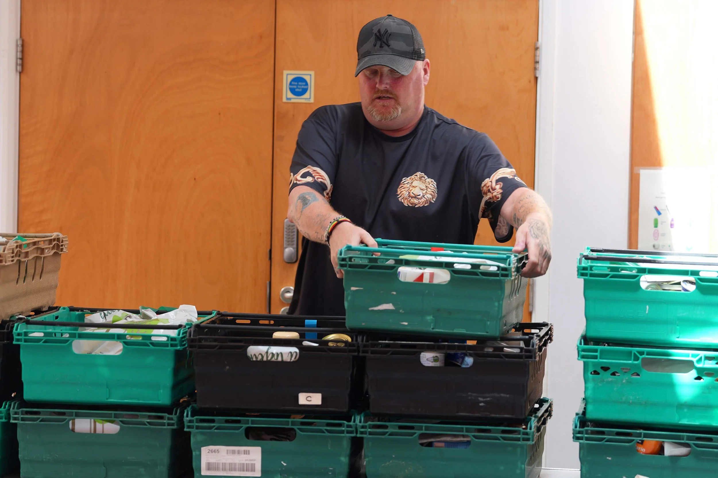 A man with tattoos on his arms, wearing a dark T-shirt and a black cap, is packing items into green plastic crates on a table - supporting the Foodbank that Grace run with the Trussell Trust. #Foodbank #Kempston