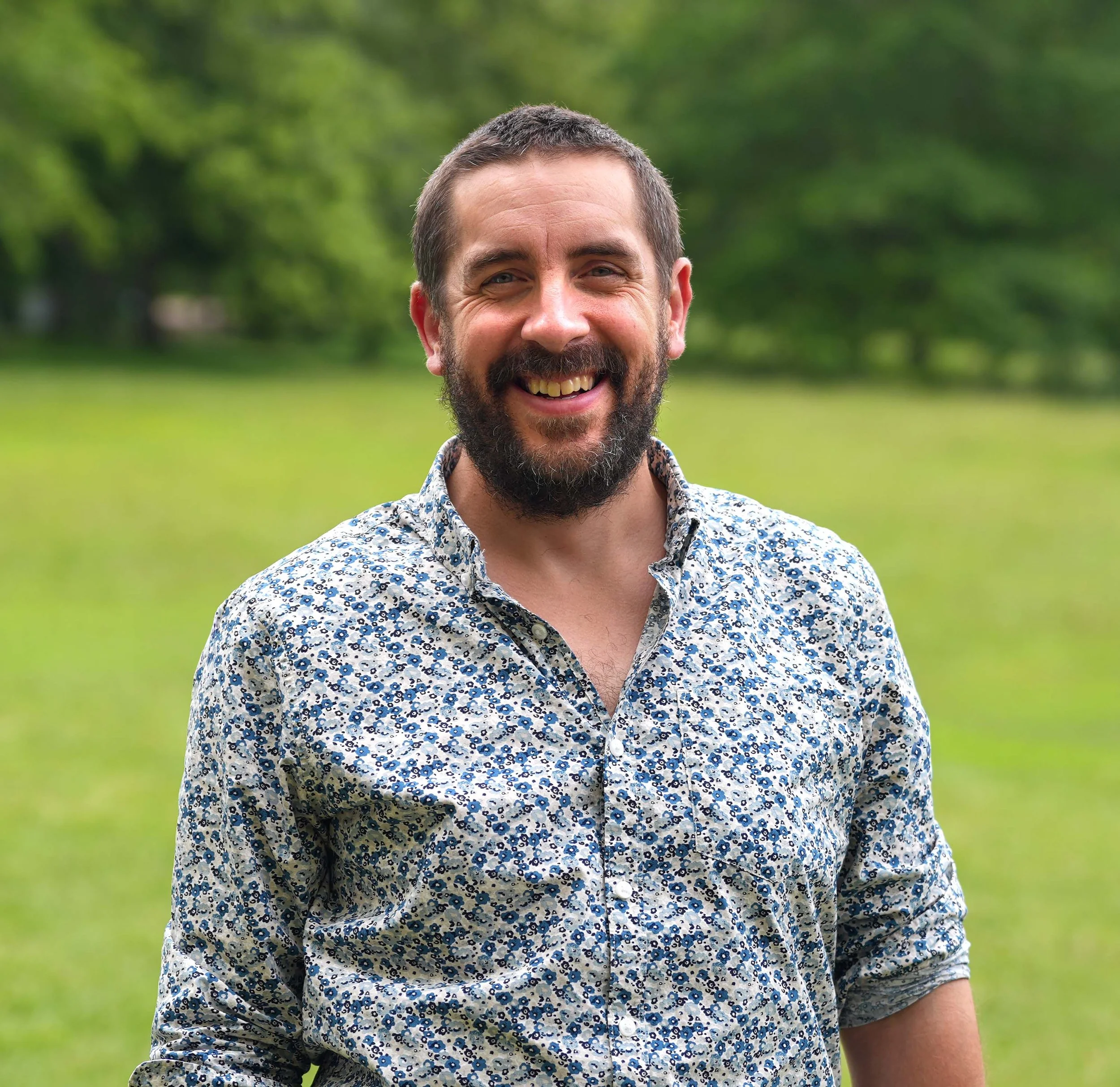 A man with short brown hair and a beard, smiling, wearing a blue and white floral patterned shirt, standing outdoors with a blurred green background.