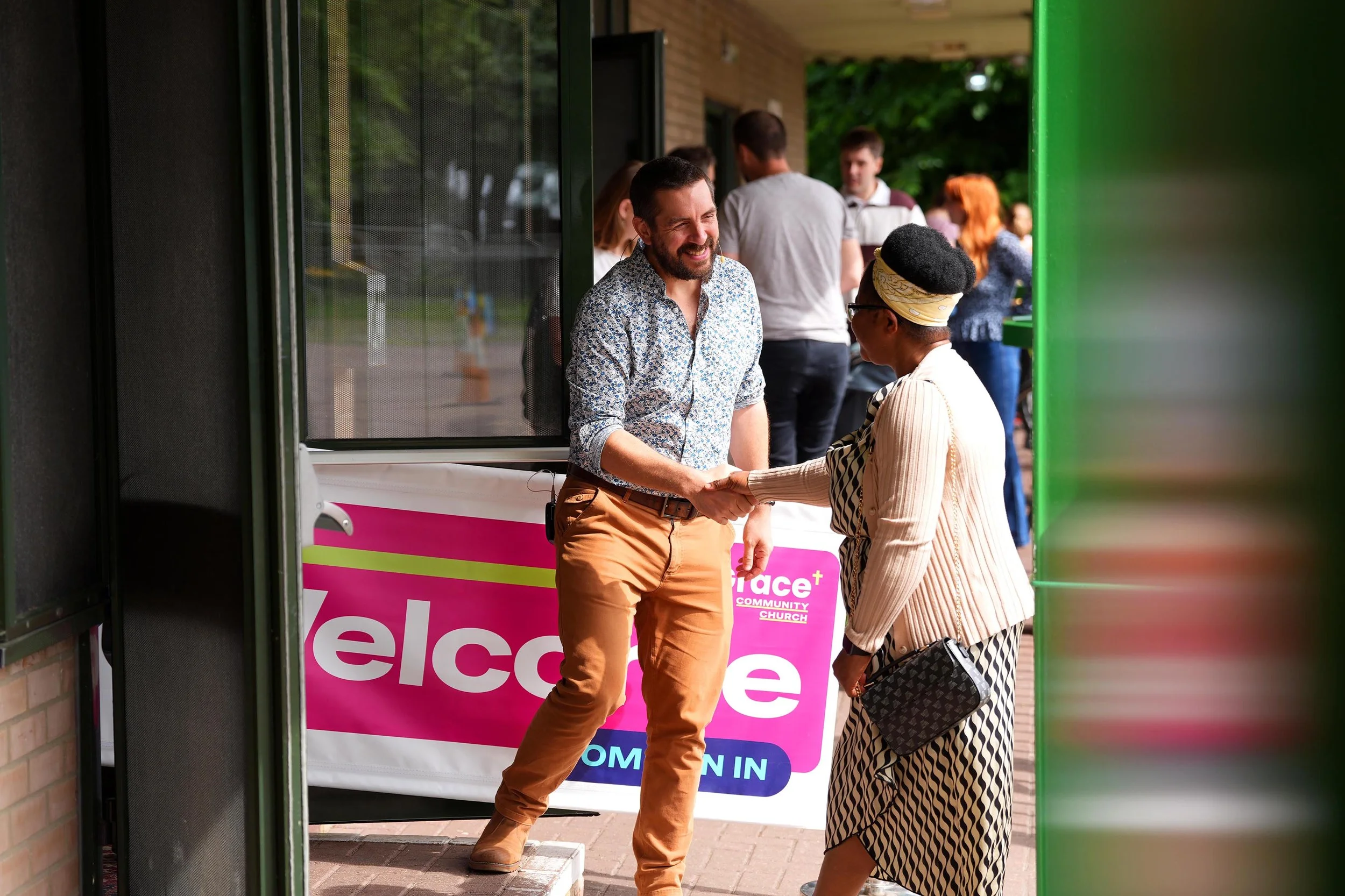 A man and a woman shaking hands outside church, with other people gathered in the background near a colorful sign that says "welcome". #Grace #Church #SundayServices #Kempston #Bedford
