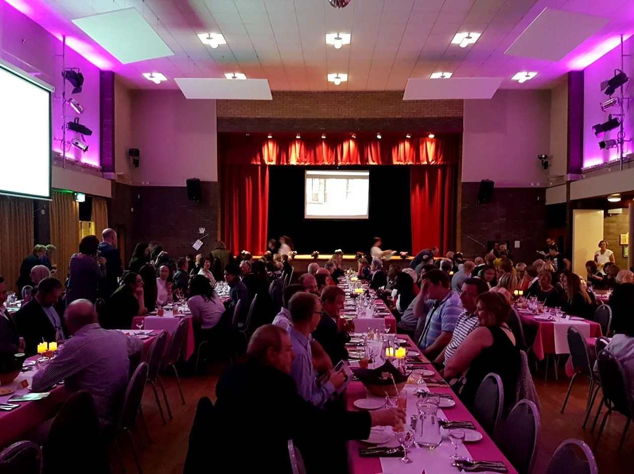 A large banquet hall filled with people sitting at long tables covered with pink tablecloths, set for a formal dinner. There is a stage with red curtains and a projection screen at the front. The room has purple and white lighting, and candles are placed on the tables.