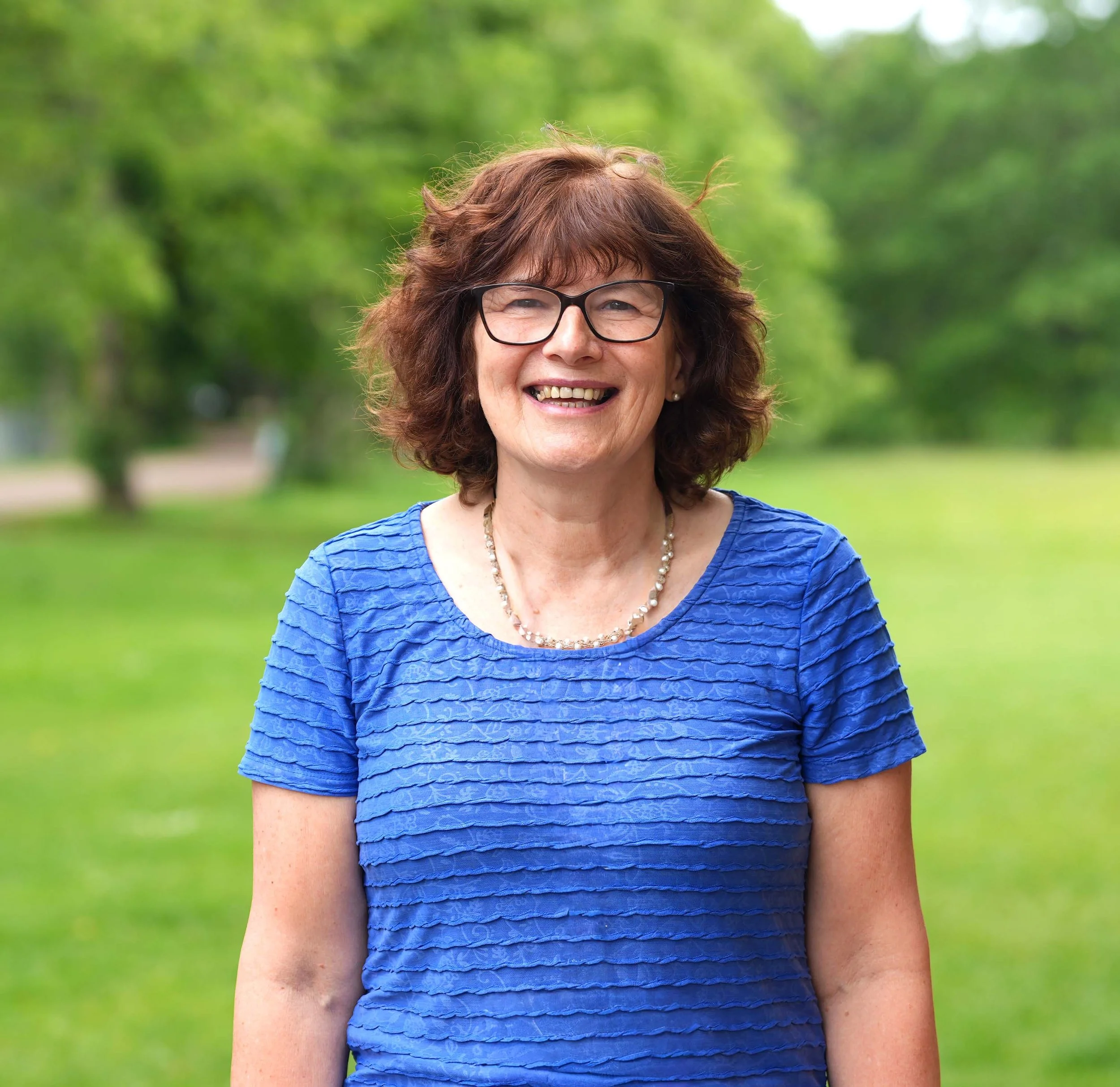 A smiling woman with glasses, brown hair, and a blue textured top standing outdoors with green trees in the background.