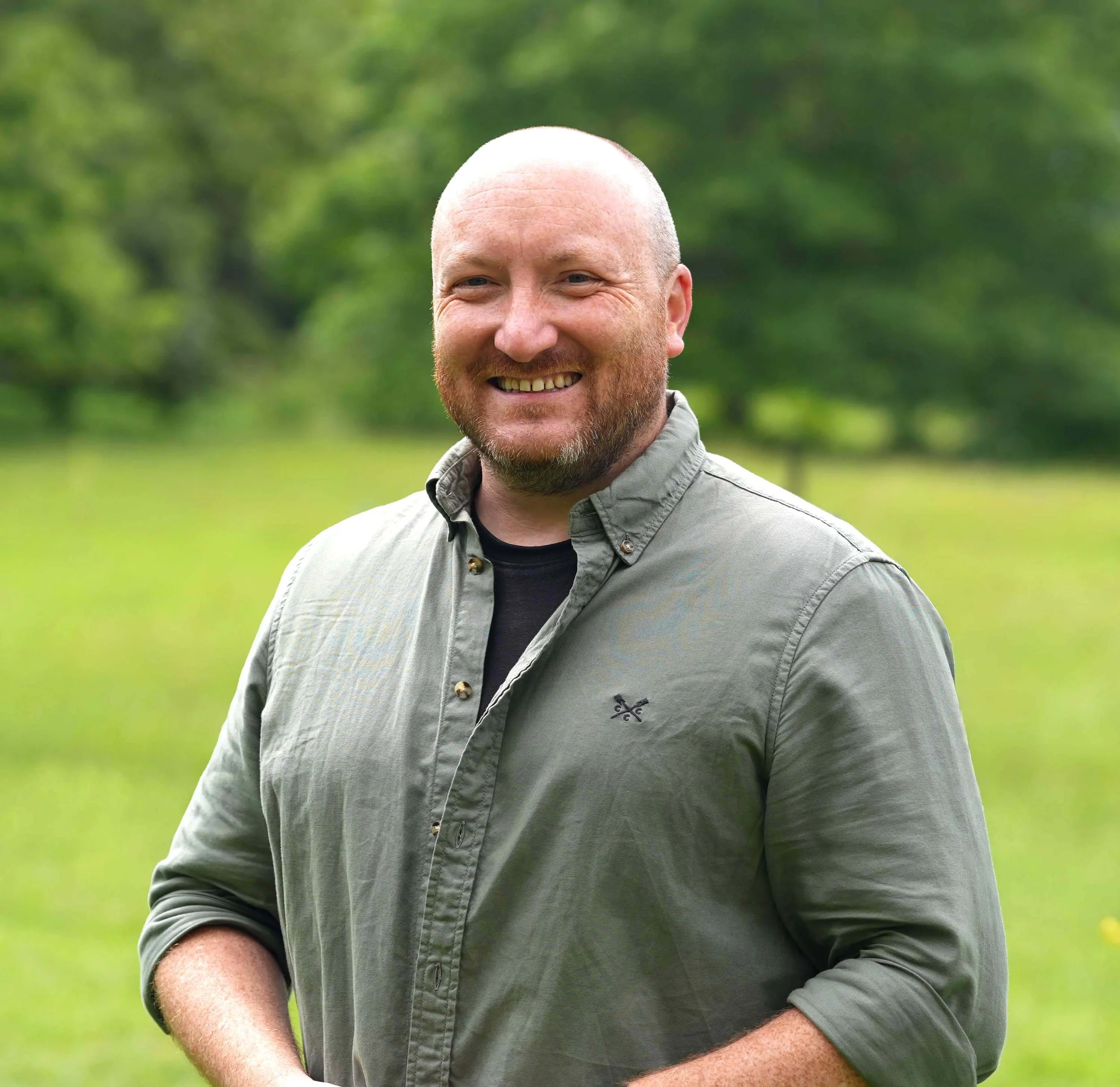 A smiling man with a beard and a shaved head standing outdoors with green blurred trees and grass in the background.