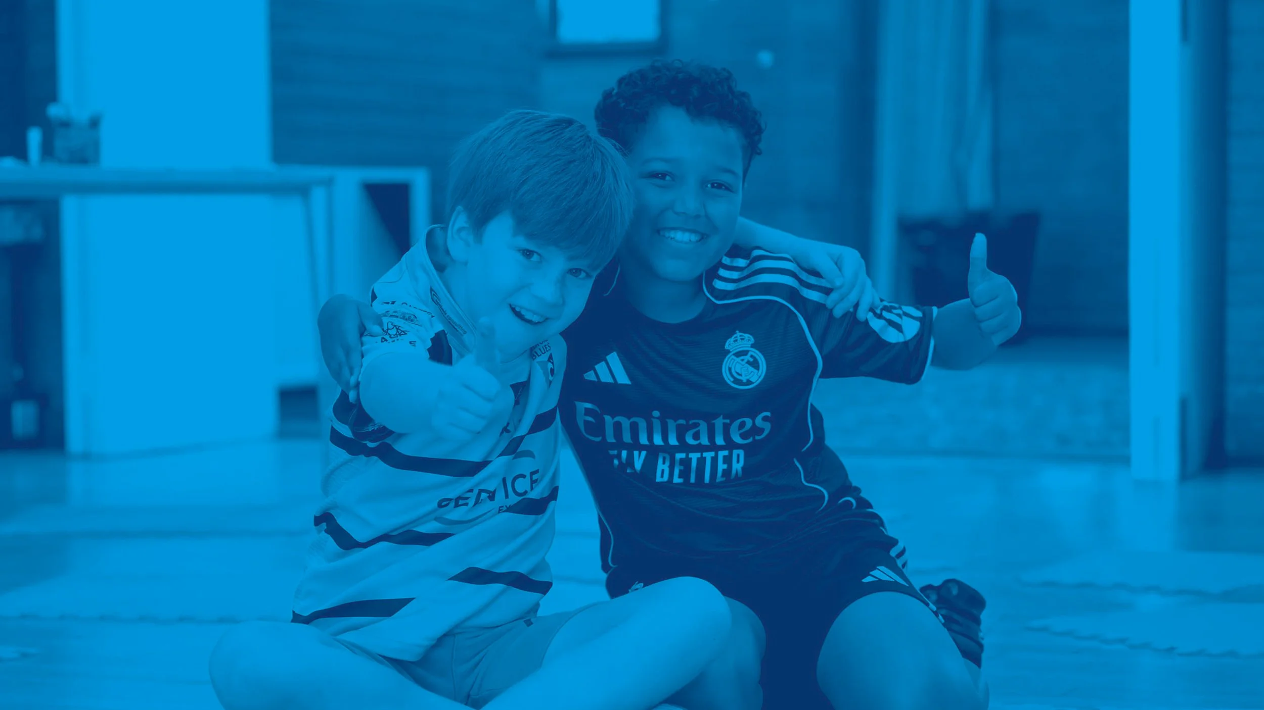 Two young boys, sitting on the floor together, smiling, with one giving a thumbs-up during one of our weekly kids clubs.  #Grace #KidsClubs