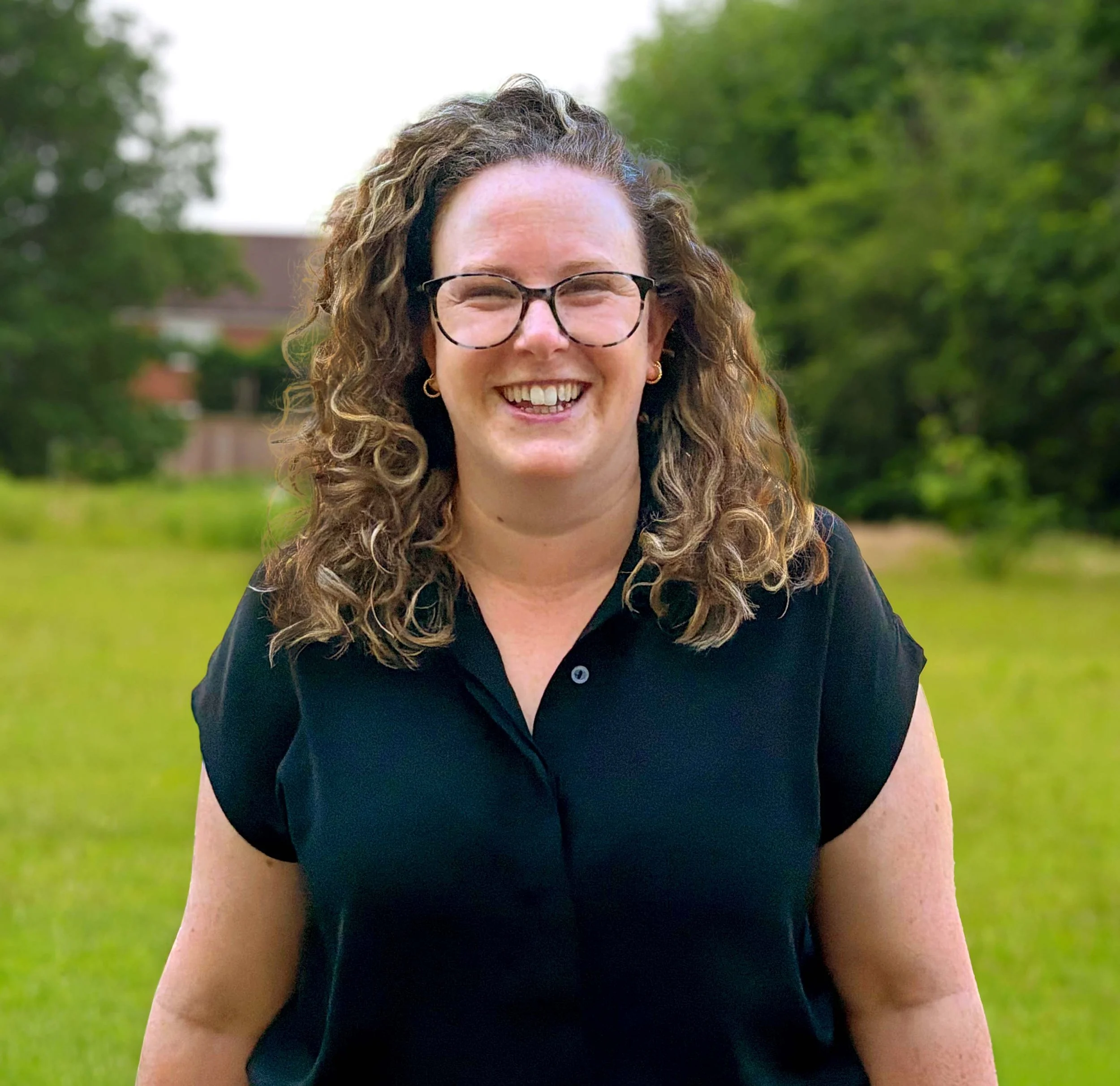A smiling young woman with curly brown hair and glasses, wearing a black shirt, outdoors with trees and a house in the background.
