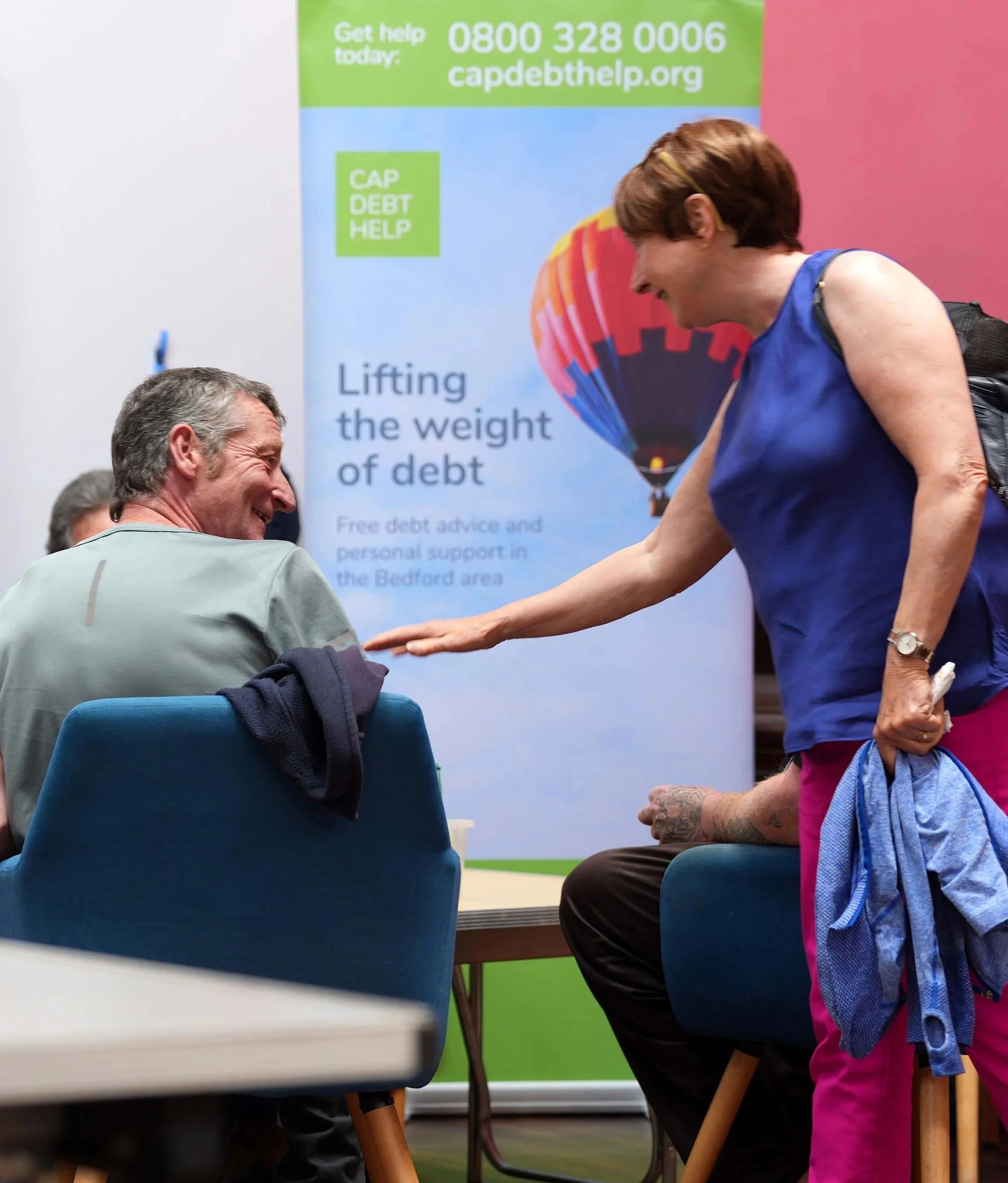 A woman smiling and reaching out her hand to a man sitting in a chair Christians Against Poverty meeting - helping 'Lifting the weight of debt' for people in our community. #Kempston #Bedford #Community #Debt