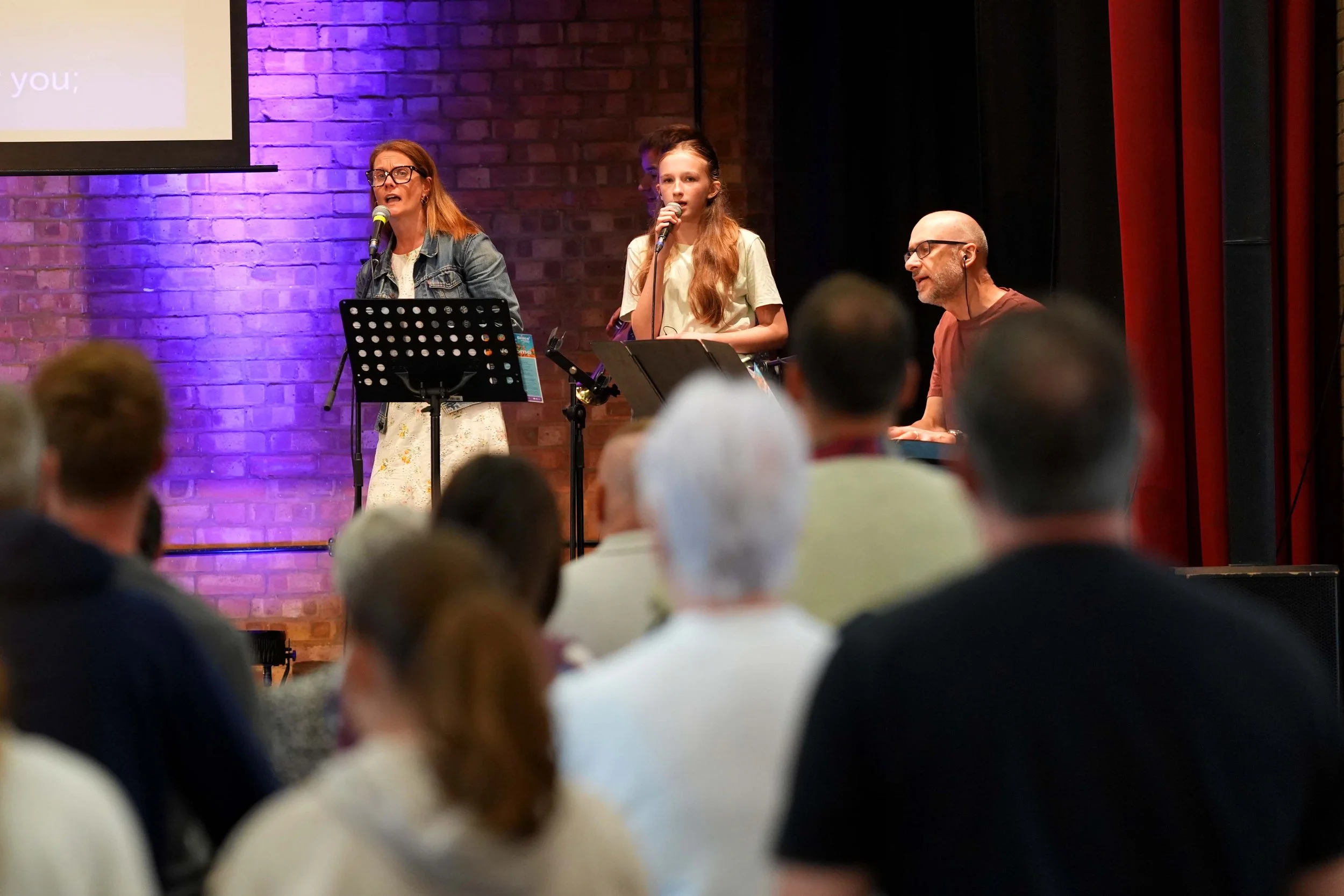 A woman with glasses and a man with glasses and a beard singing into microphones on stage during a performance, with an audience in the foreground.