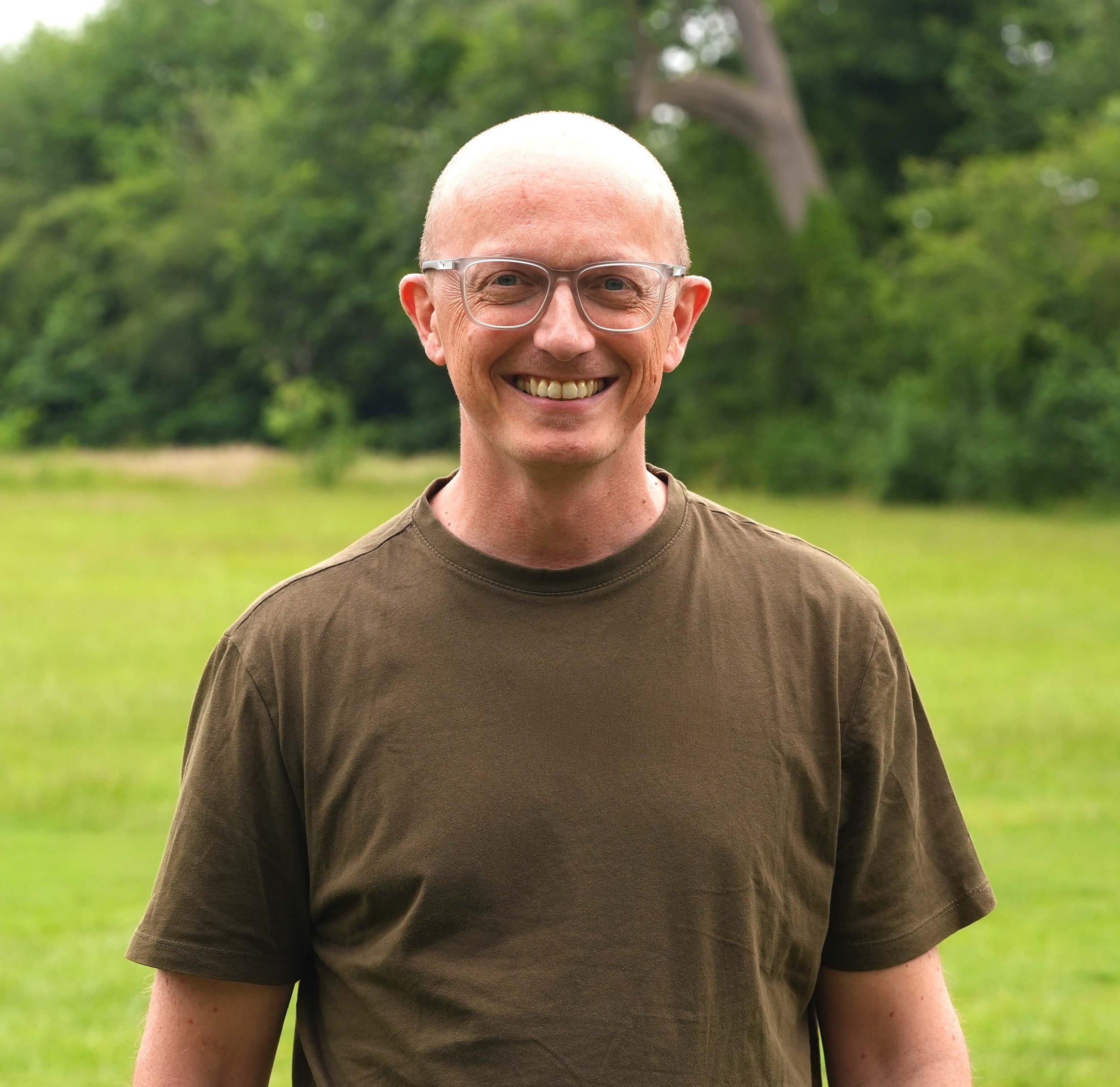 Close-up of a smiling man with glasses and a shaved head standing outdoors with green trees in the background.