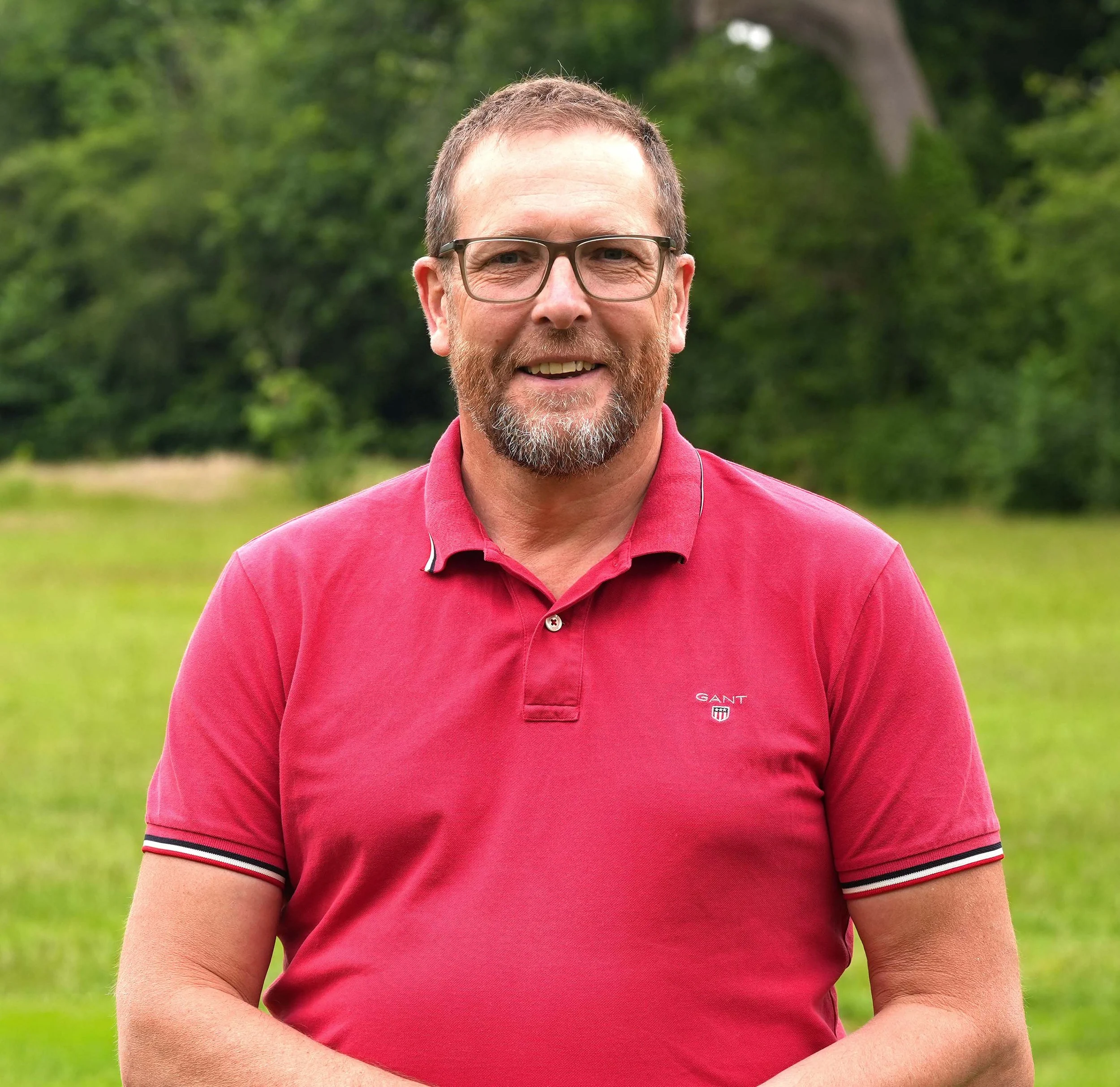 Close-up of a smiling middle-aged man with glasses, short brown hair, and facial hair, wearing a pink polo shirt, standing outdoors with green trees and grass in the background.