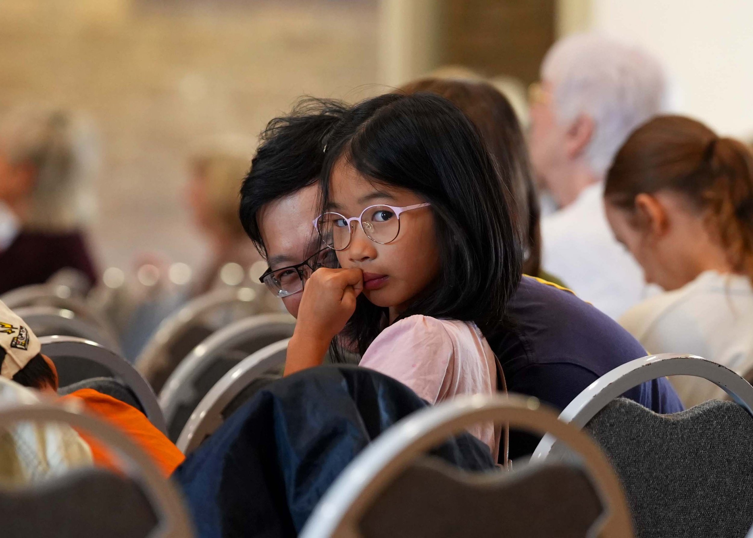 A young girl with glasses and long black hair is resting her chin on her hand and looking directly at the camera. She is sitting amidst a crowd of people after a Sunday Service at Grace Community Church, Bedford.