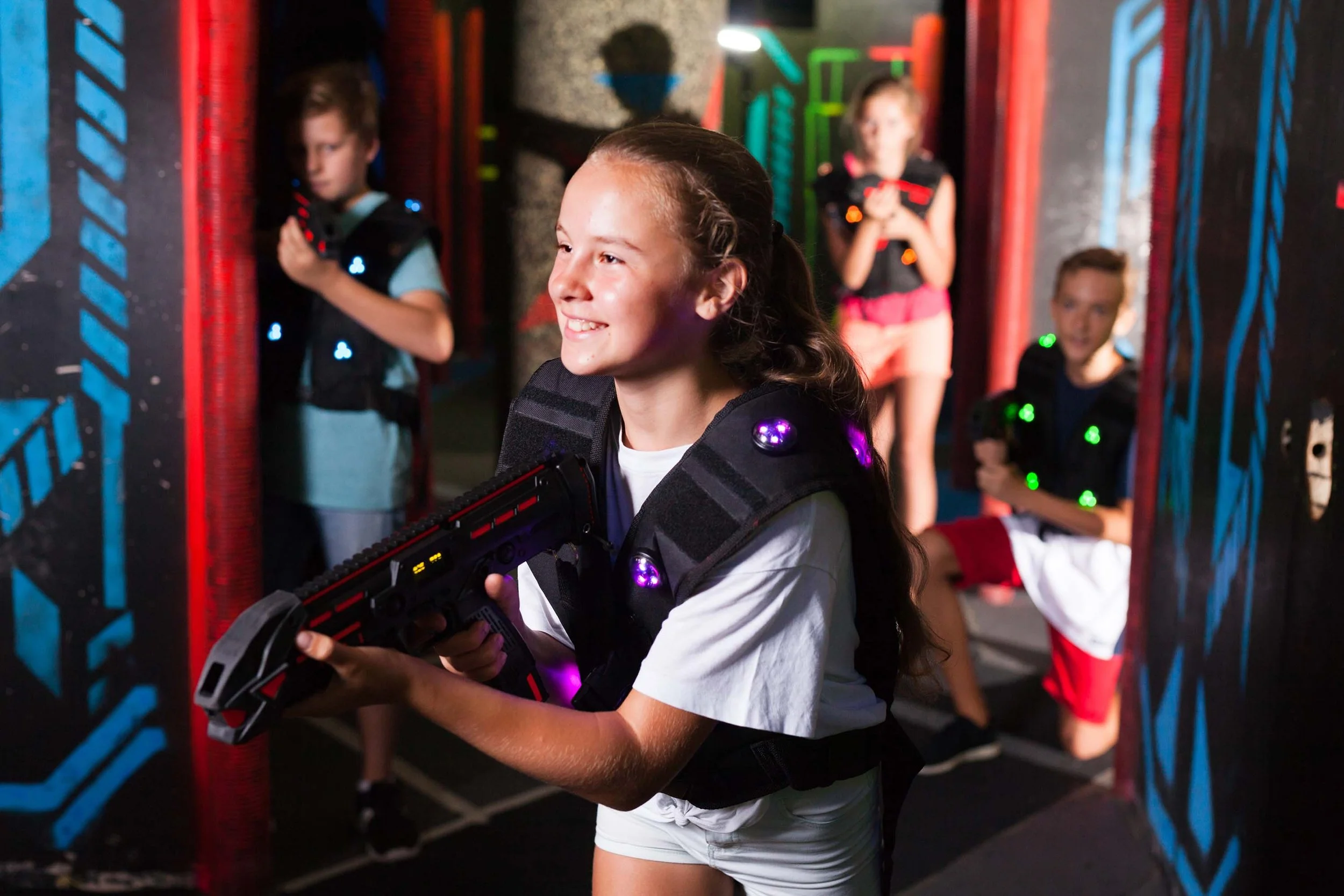 Children playing laser tag game, with one girl smiling and holding a laser gun, wearing a vest with glowing lights, in an indoor laser tag arena.