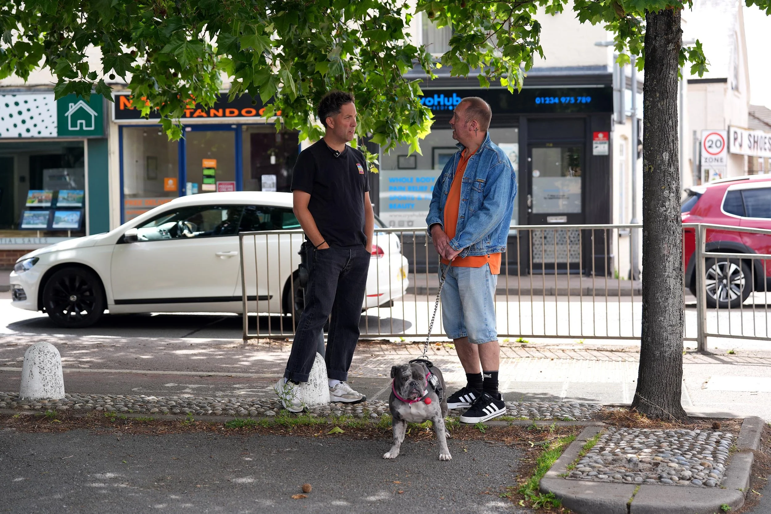 Two men, one in a black shirt and the other in a denim jacket and orange shirt, are standing and talking on the sidewalk under a tree with green leaves. They talk about life, faith, family.