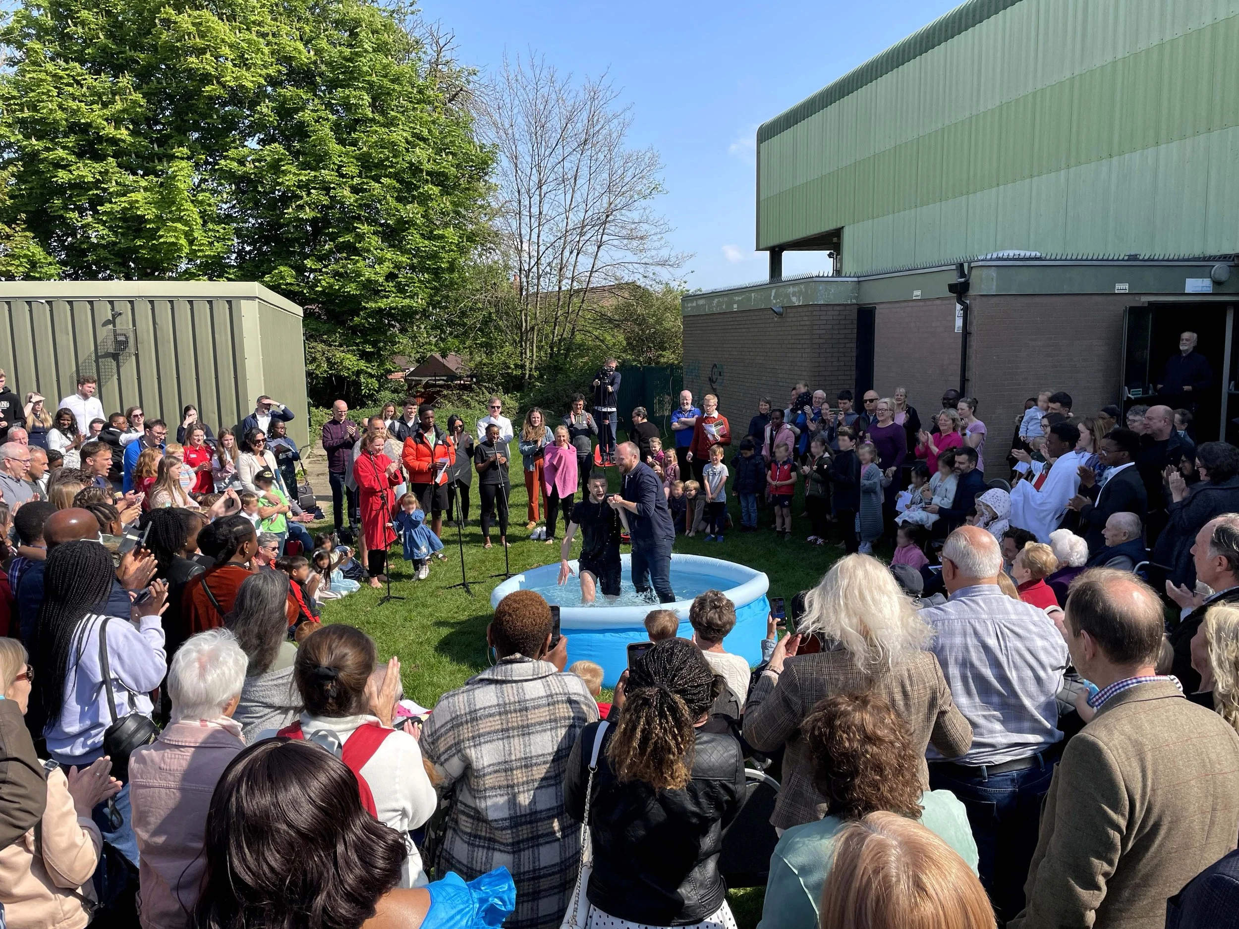 A diverse crowd of people gathered outdoors on a sunny day, gathered round a small pool where Christian baptisms are taking place.