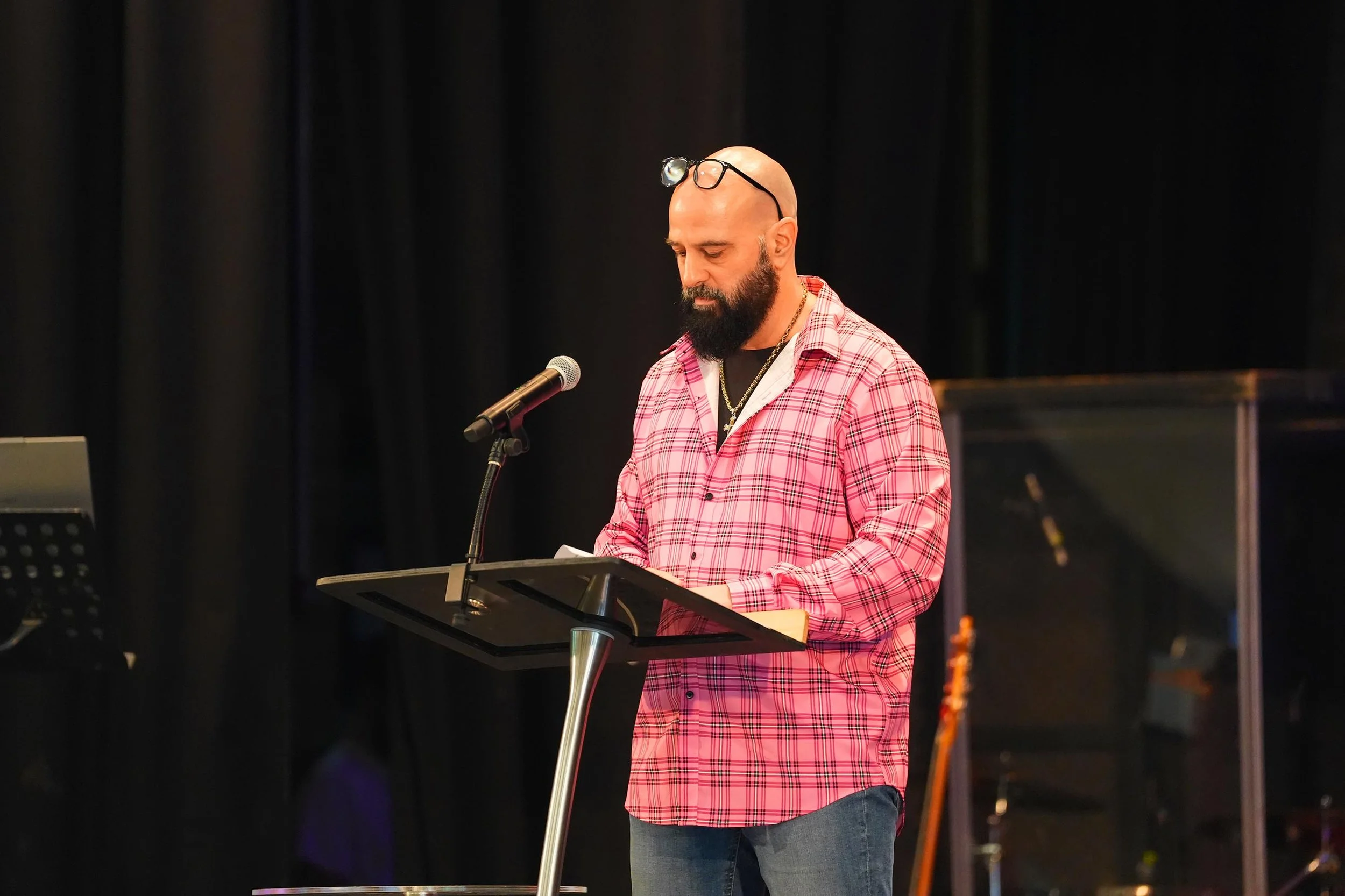 A man with a bald head and a beard, wearing glasses on his head, a pink and white checkered shirt, and a necklace with a cross, reading from a script at a lectern with a microphone in a dimly lit setting.
