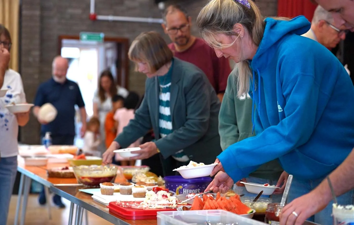 People serving themselves food at a buffet table filled with various dishes like watermelon, cupcakes, and salads at Big Sunday Lunch at Grace
