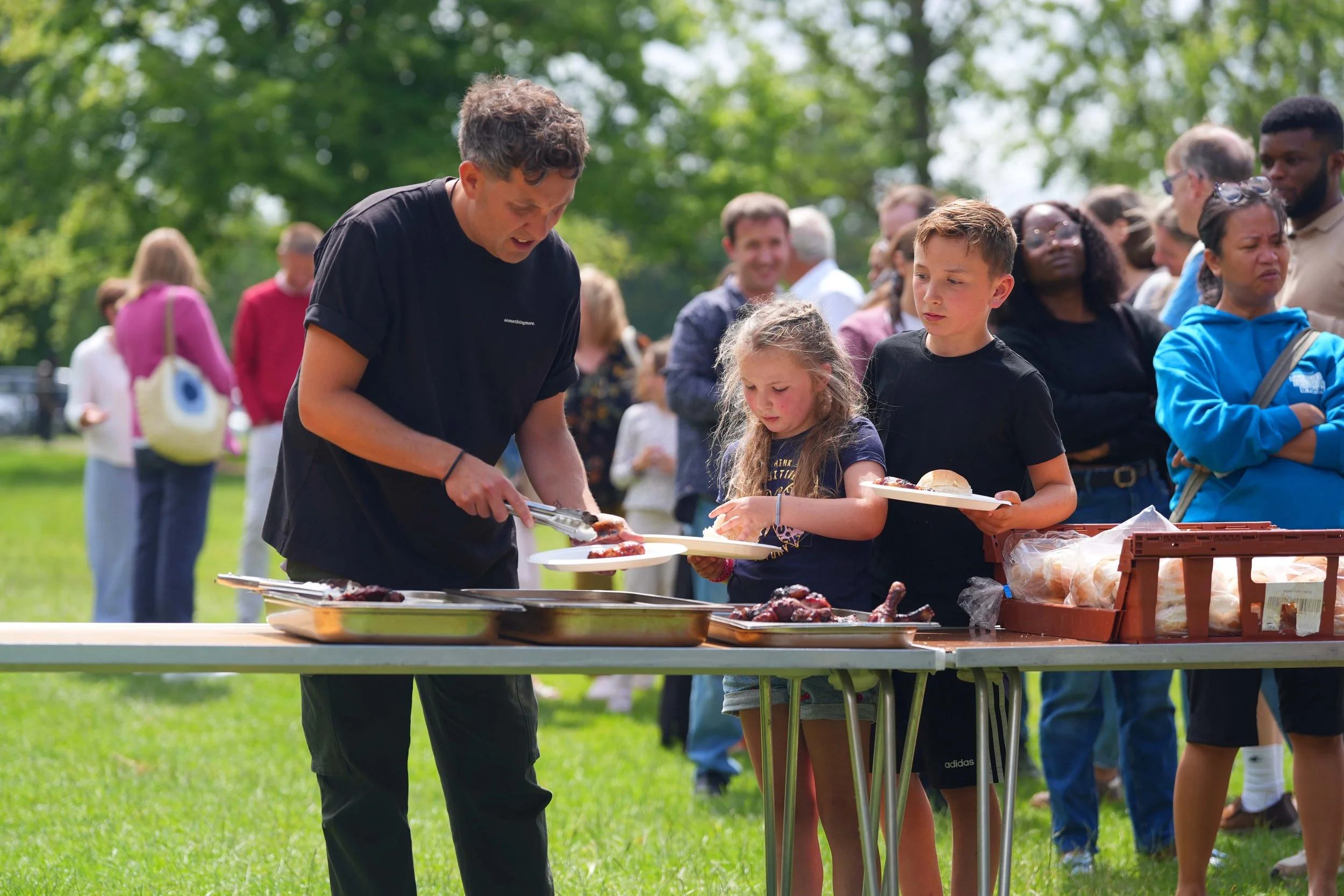 People at an outdoor gathering serving food at a buffet table with grilling items, in a park with green trees. A Grace family lunch.