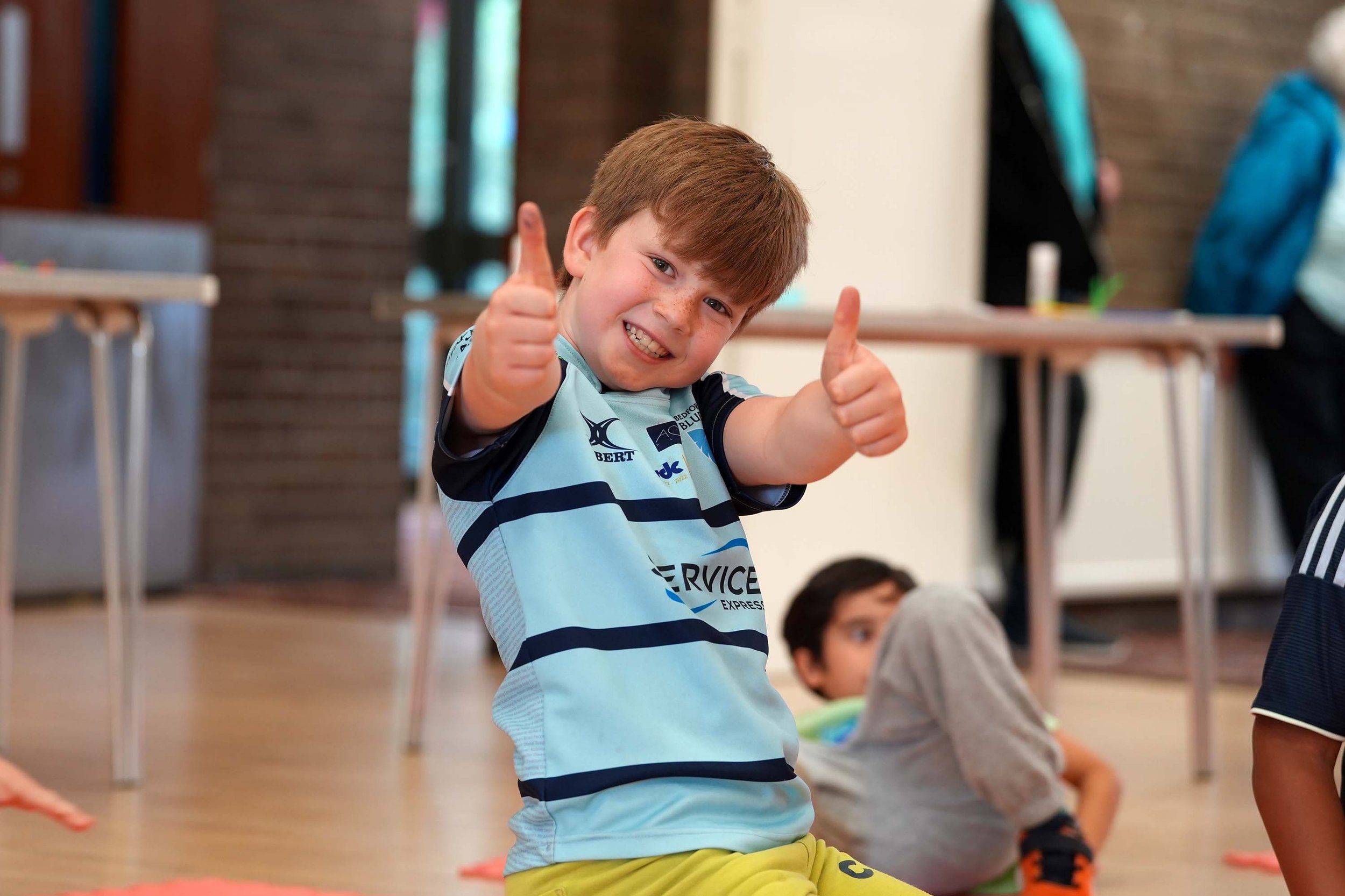 A young boy with brown hair giving a thumbs-up while kneeling, with other children sitting on the floor in the background.