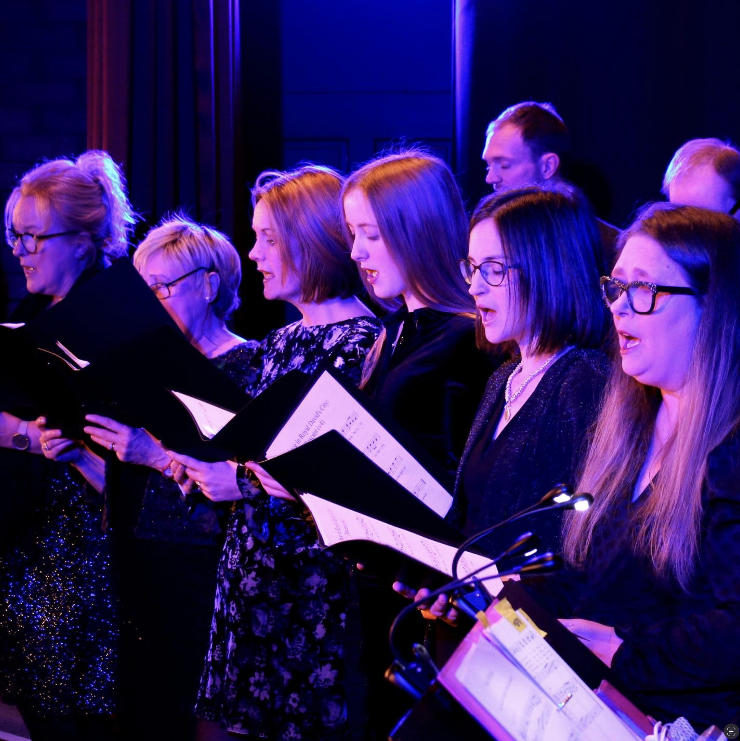 A group of choir singers performing on stage with sheet music, illuminated by blue and purple stage lighting.