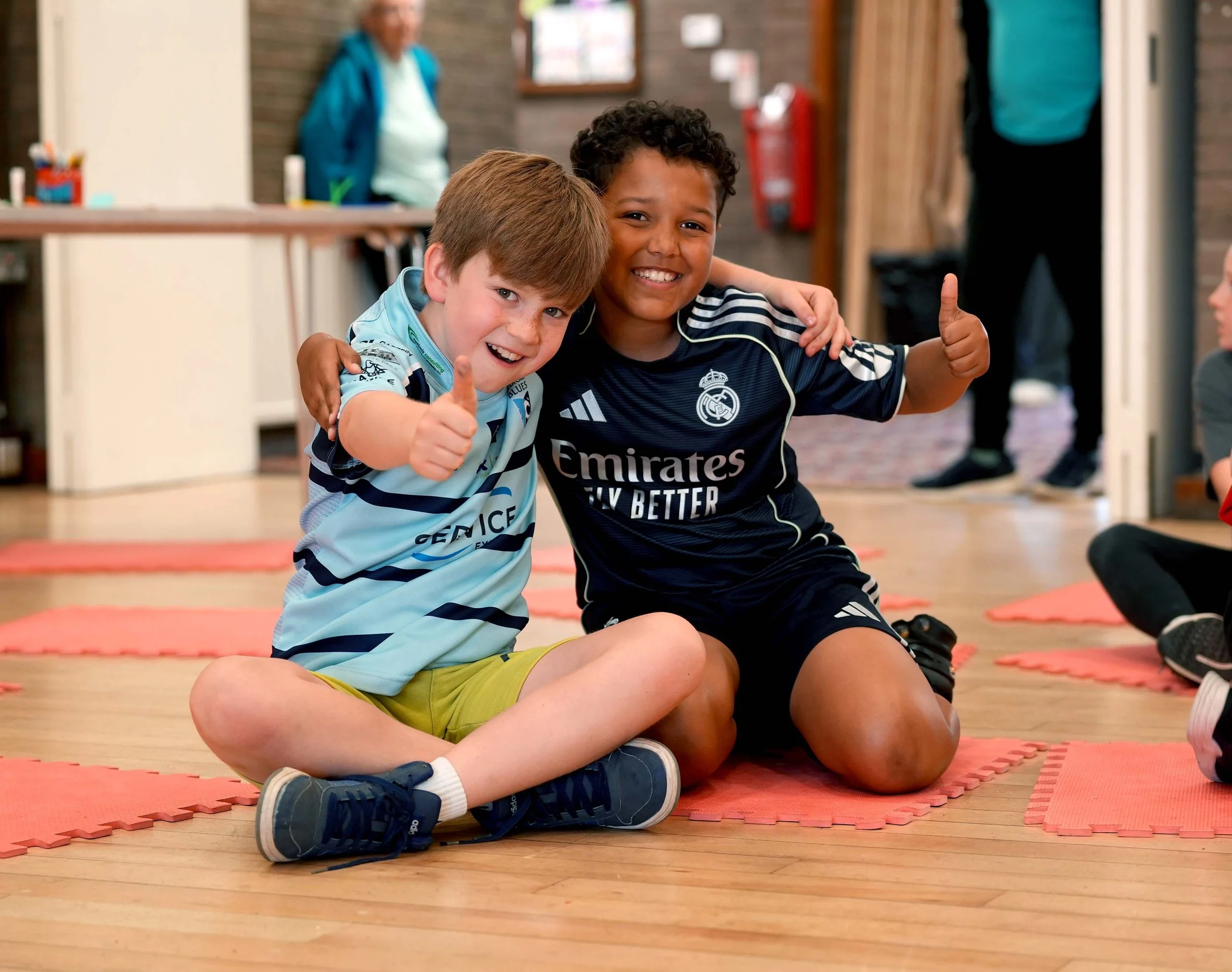Two young boys sitting on a red foam mat, smiling and giving thumbs up at a kids club in Kempston