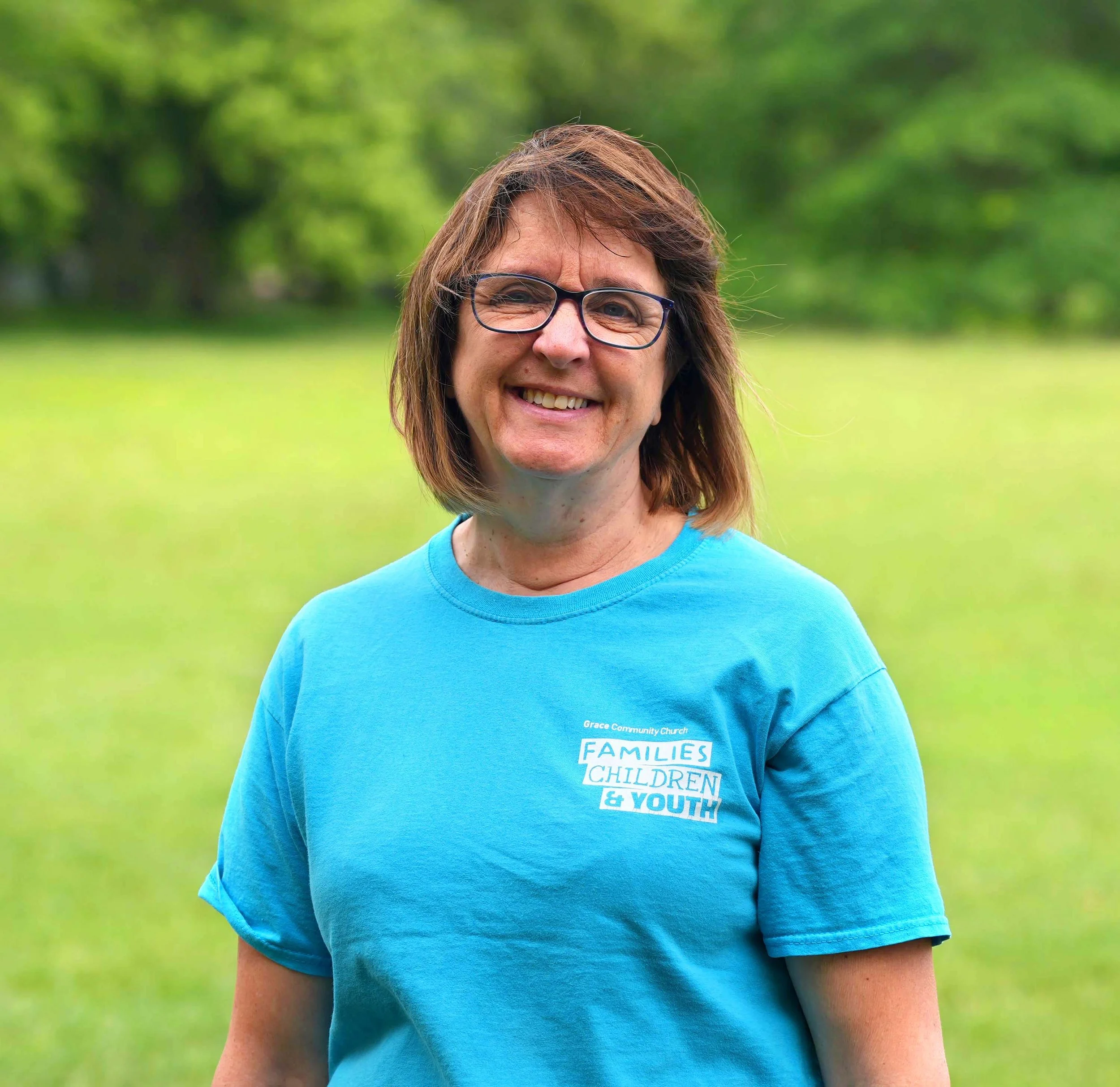 Close-up of a smiling woman with glasses and shoulder-length brown hair, wearing a blue T-shirt outdoors with a blurred green background.