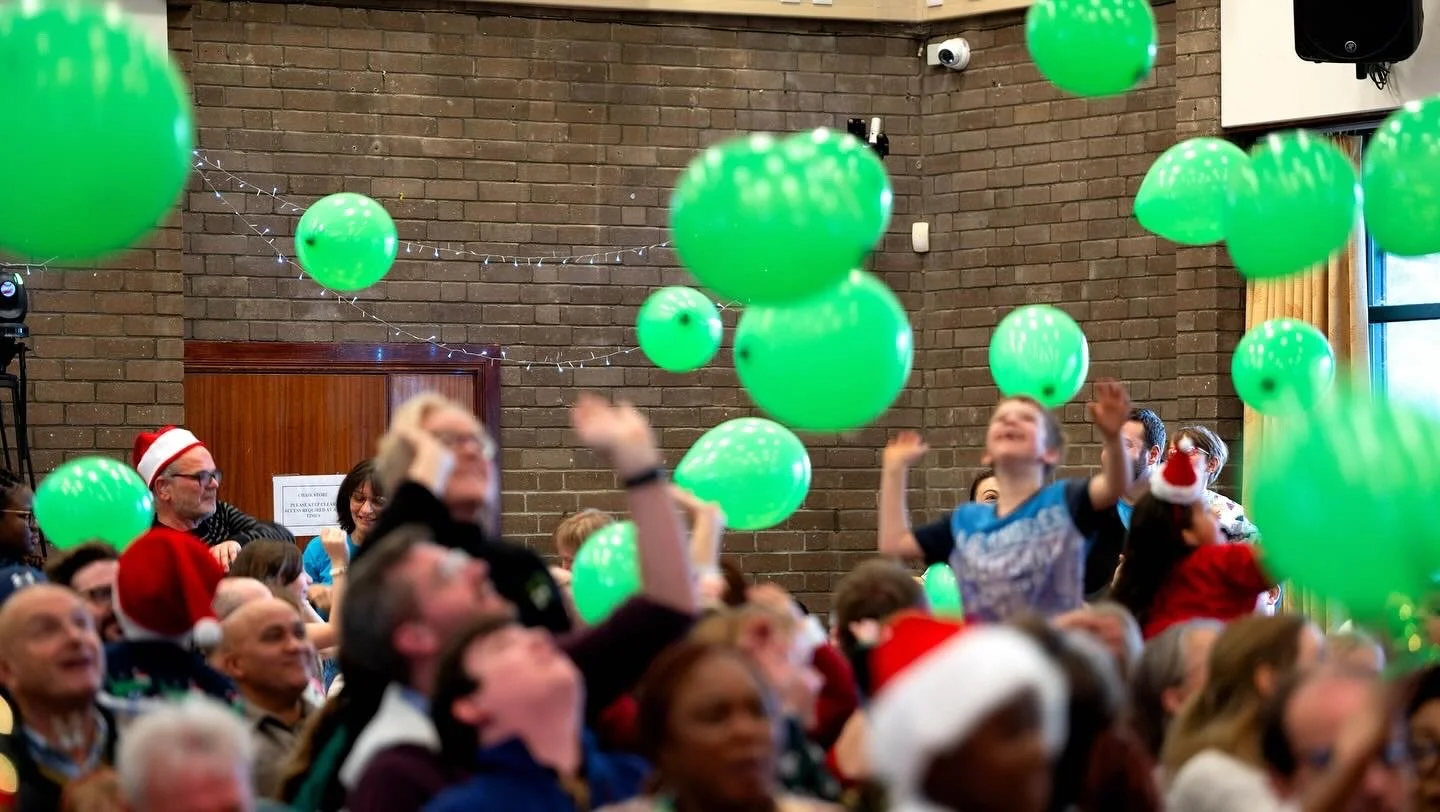 People celebrating at a festive event with many green balloons, some wearing Santa hats and Christmas accessories, in a decorated indoor venue.