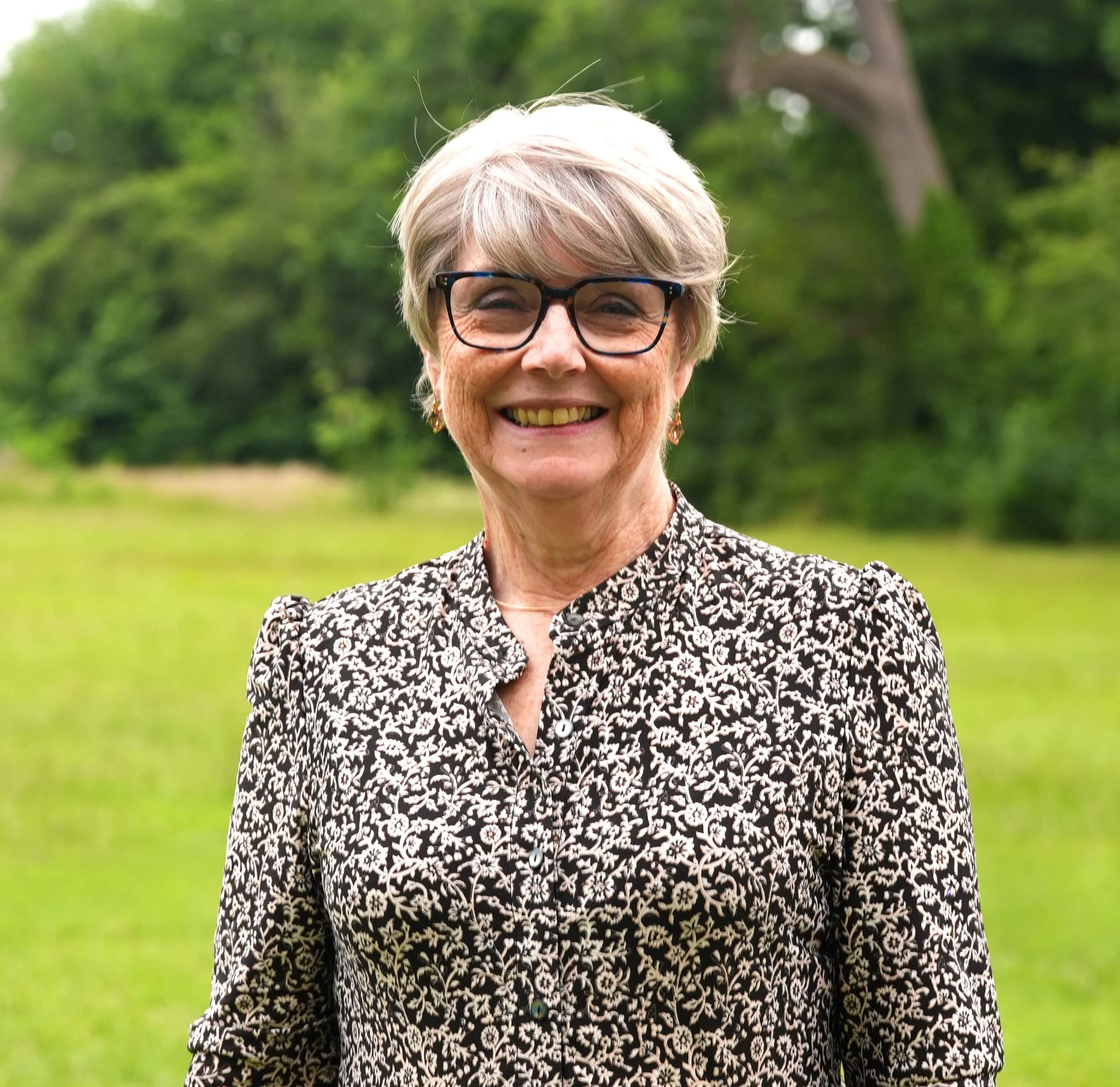 A smiling elderly woman with short gray hair, wearing glasses, standing outdoors in front of green trees, dressed in a black and white patterned blouse.
