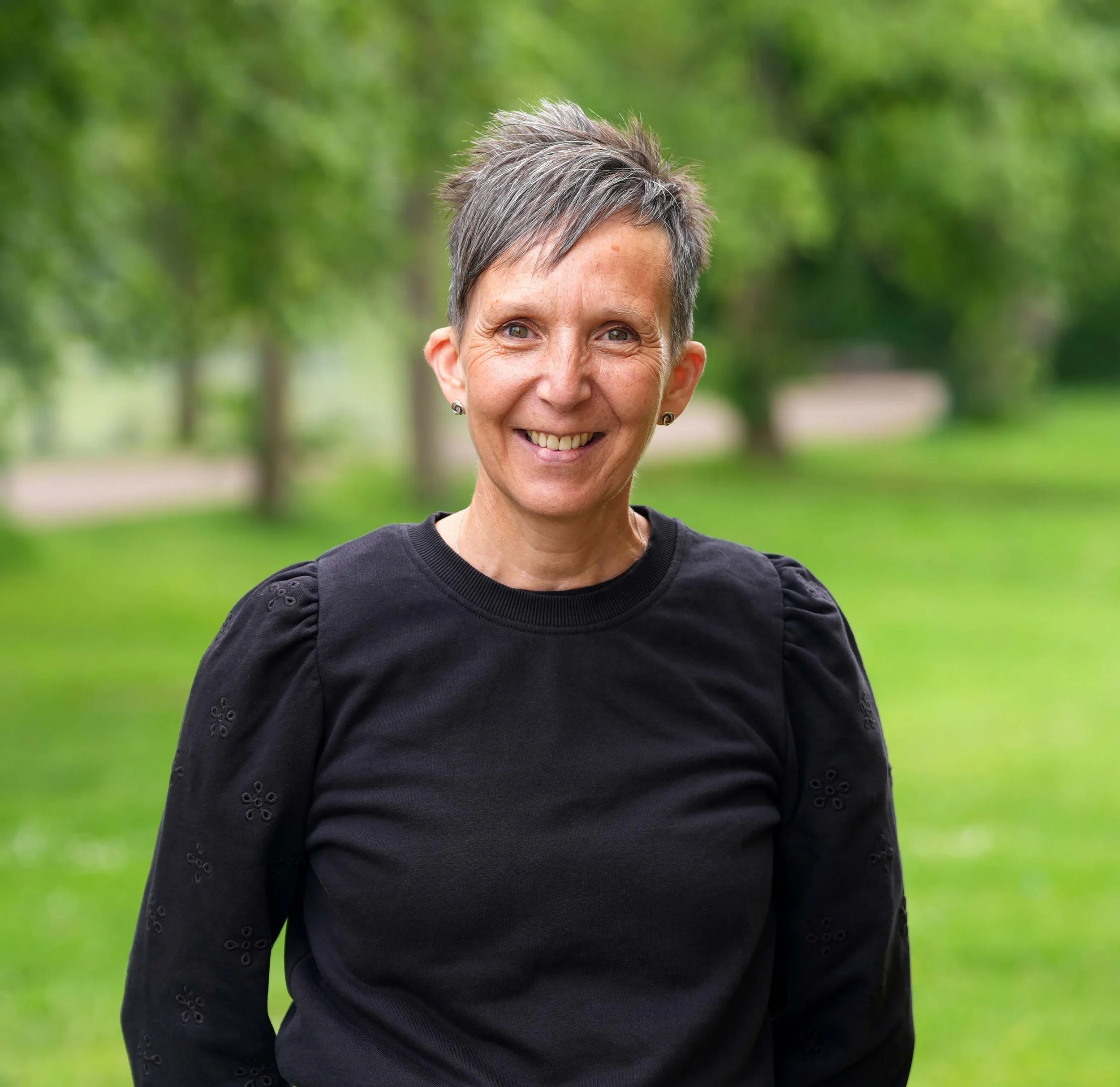 A smiling woman with short gray hair wearing a black top with embroidered details, standing outdoors with green trees in the background.