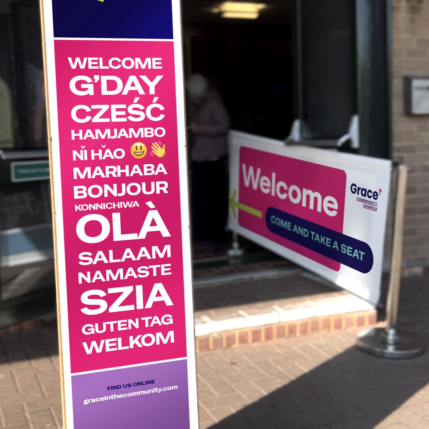 A tall pink and purple welcome sign with translations of the word 'welcome' in multiple languages, and a smaller white sign with additional writing, outside the Addison Centre, in Kempston where Grace Community Church meet each week.