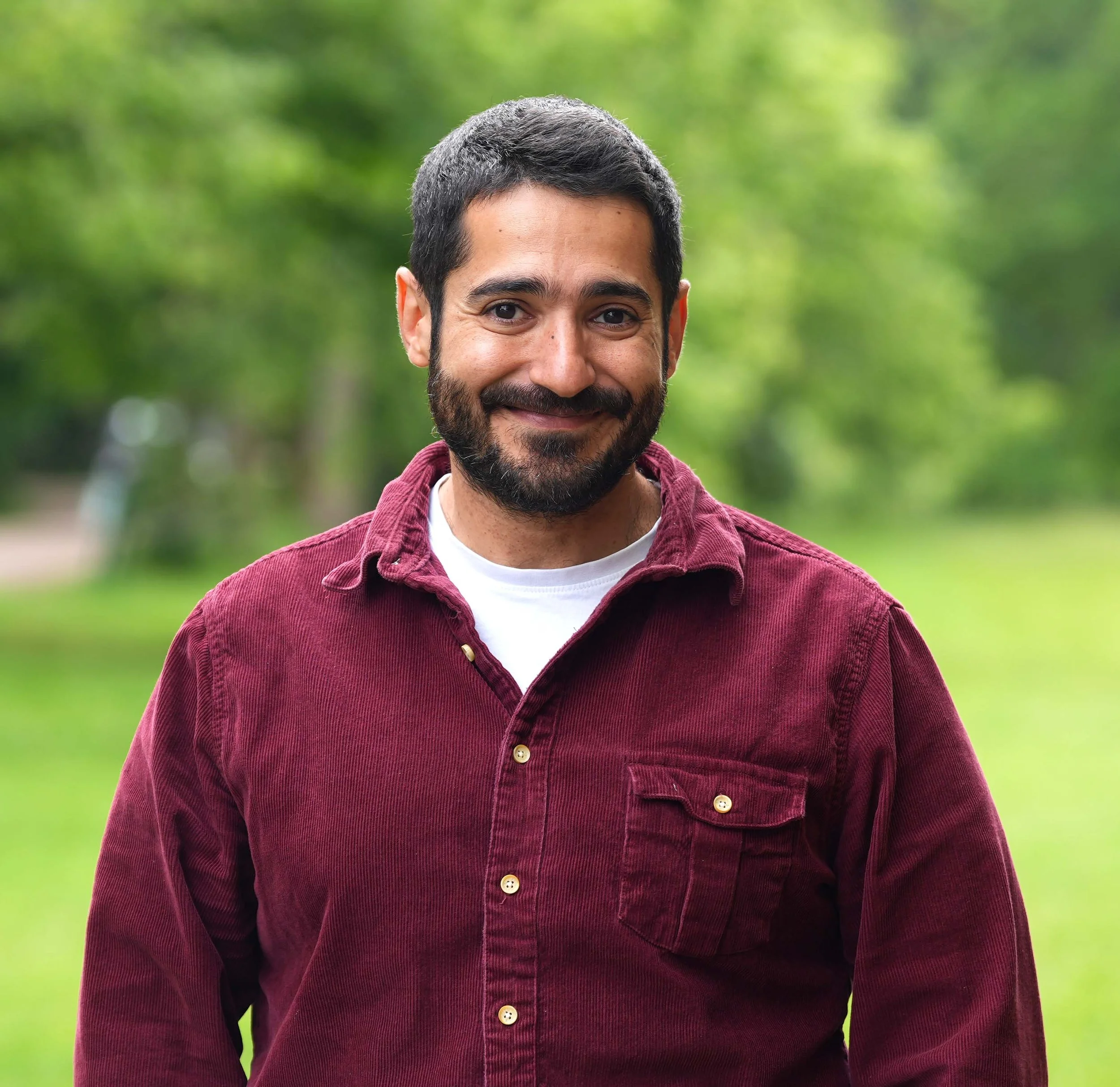 A smiling man with dark hair, a beard, and mustache wearing a red jacket outdoors with blurred green trees in the background.