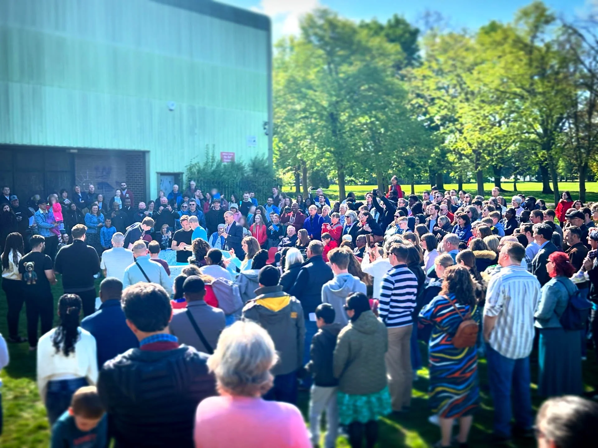 A large outdoor gathering of diverse people, including adults and children, participating in a Baptism in Addison Howard Park in Kempston. 