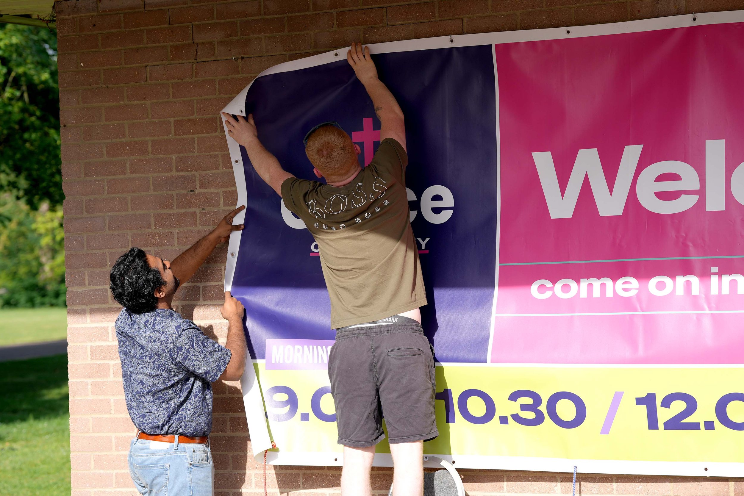 Two volunteers are hanging a colorful welcome banner at the Addison Centre for a Grace Sunday Service