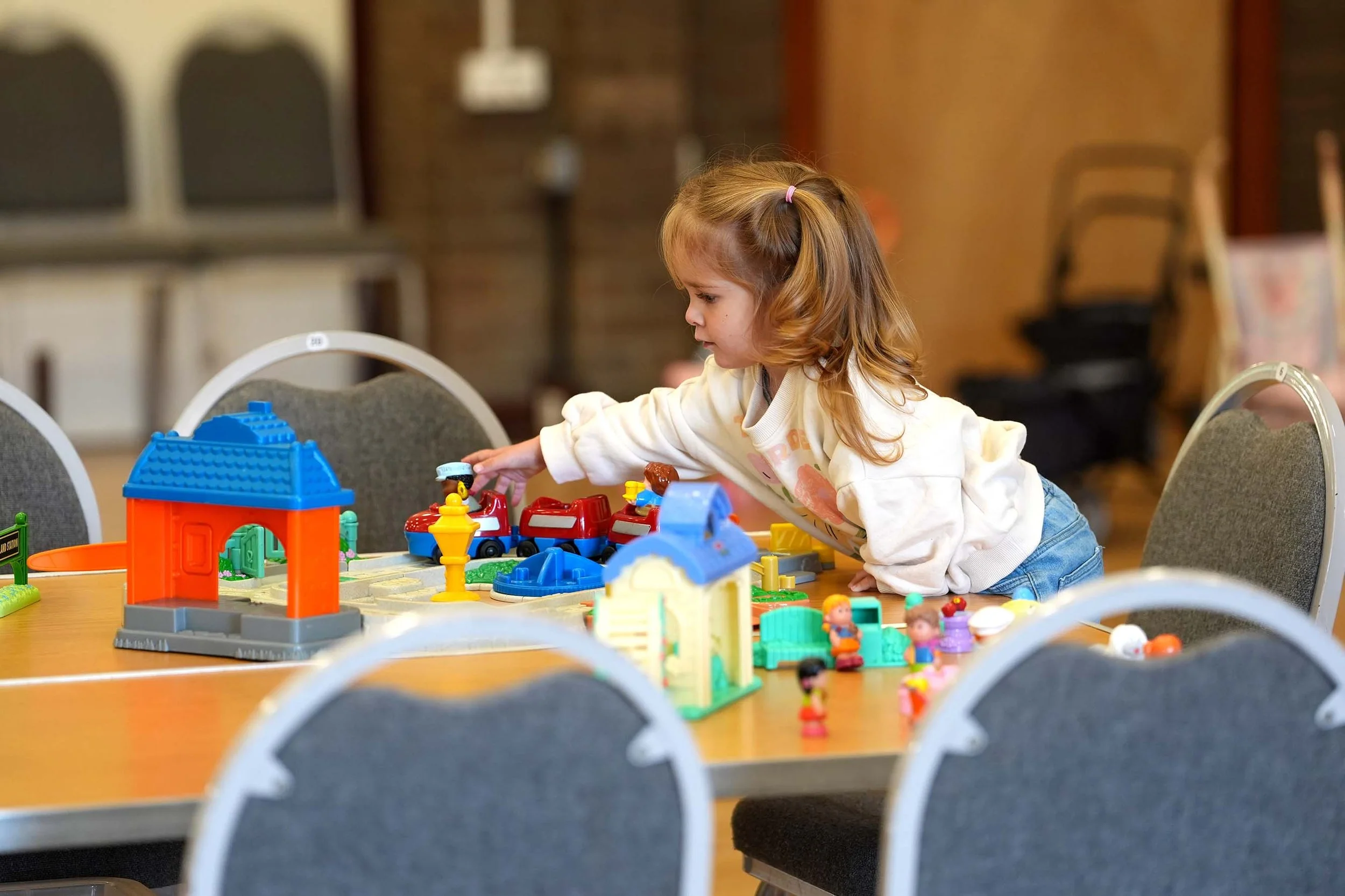 A young girl with light brown hair in a ponytail wearing a white sweatshirt is playing with colorful LEGO sets on a table in a room with chairs and a brick wall in the background.