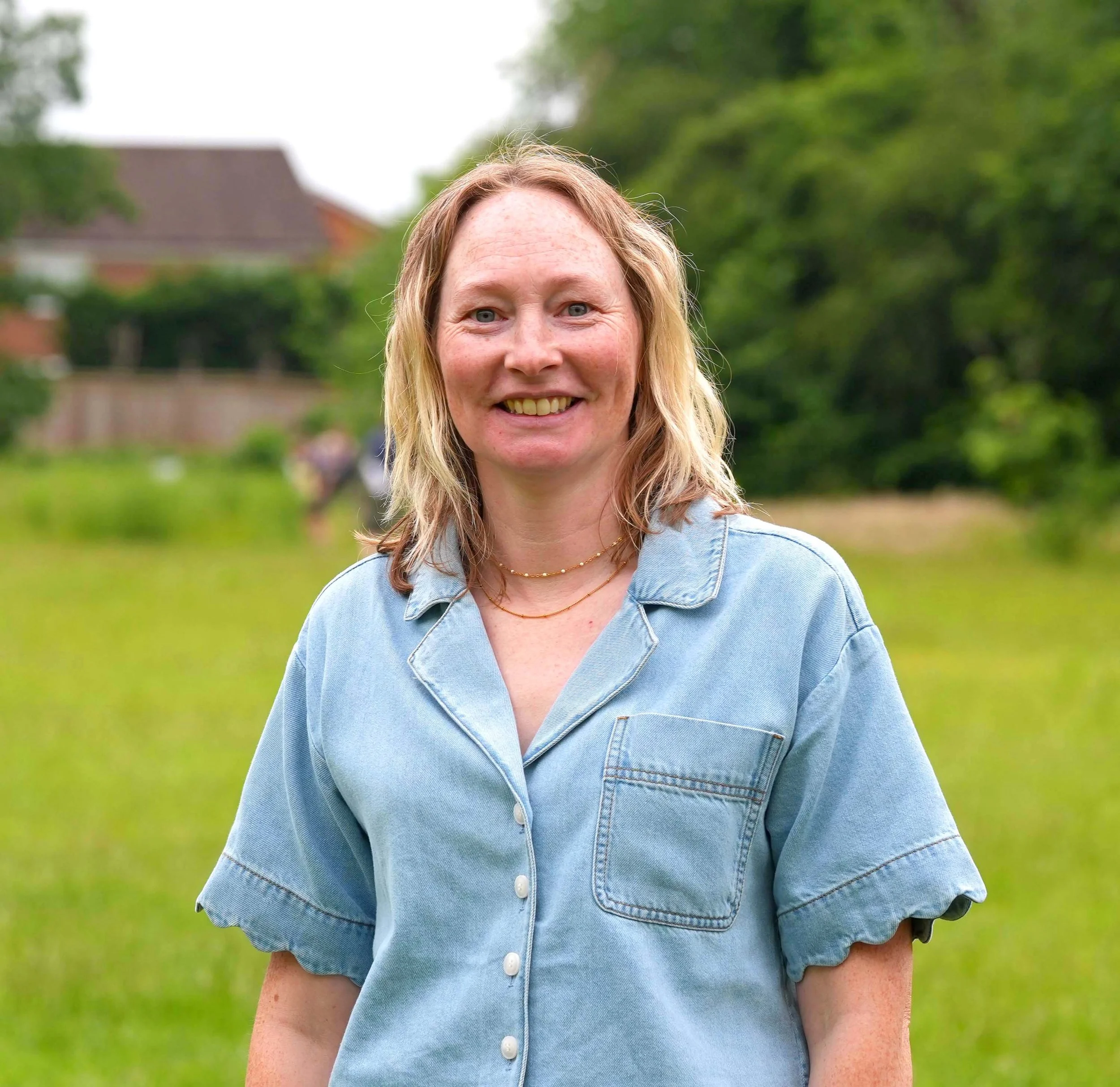 A woman with blonde hair smiling outdoors wearing a light denim button-up shirt, with green trees and a house in the background.