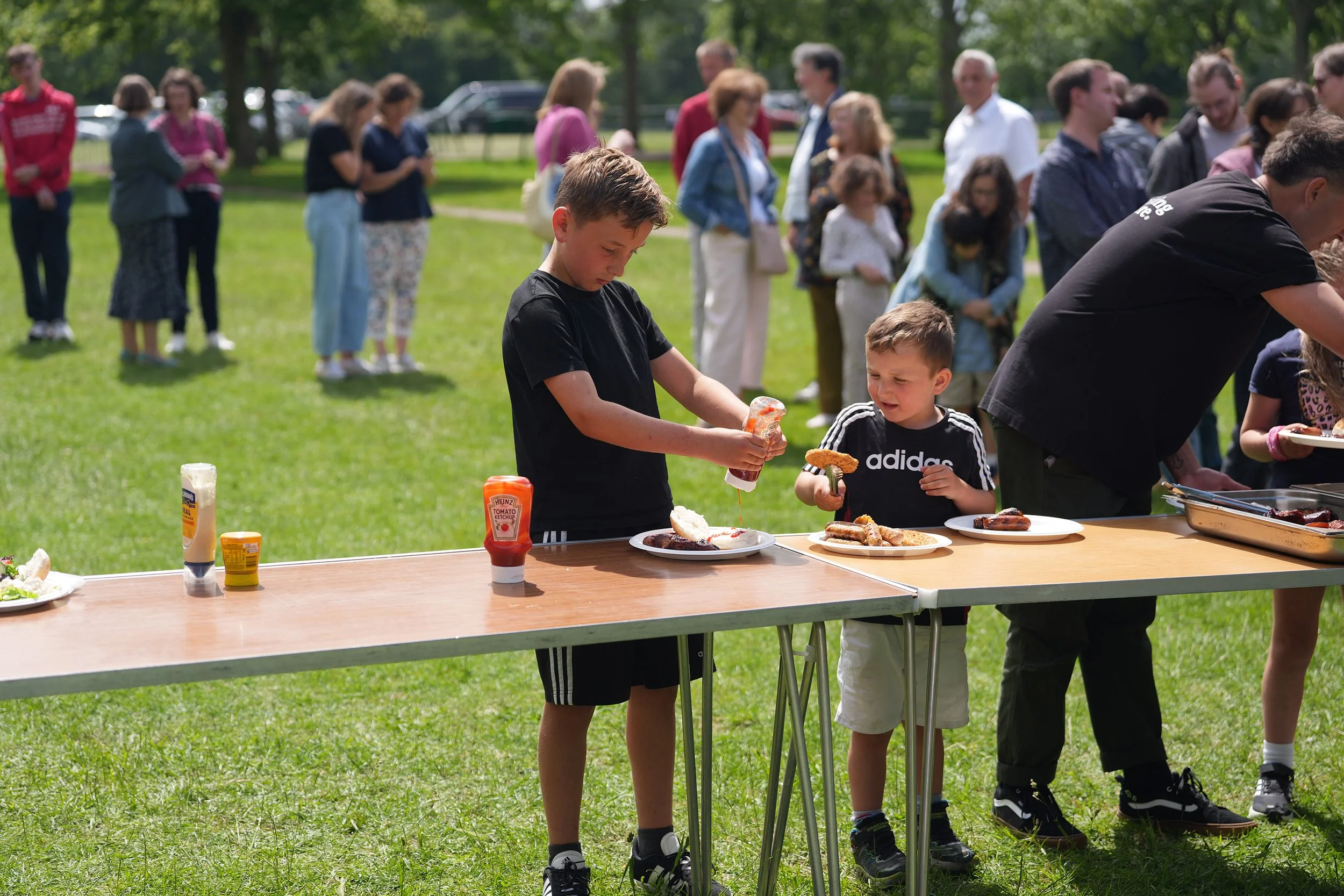 Children and adults gather at an outdoor picnic table with hot dogs, buns, and condiments during a sunny day in a park.