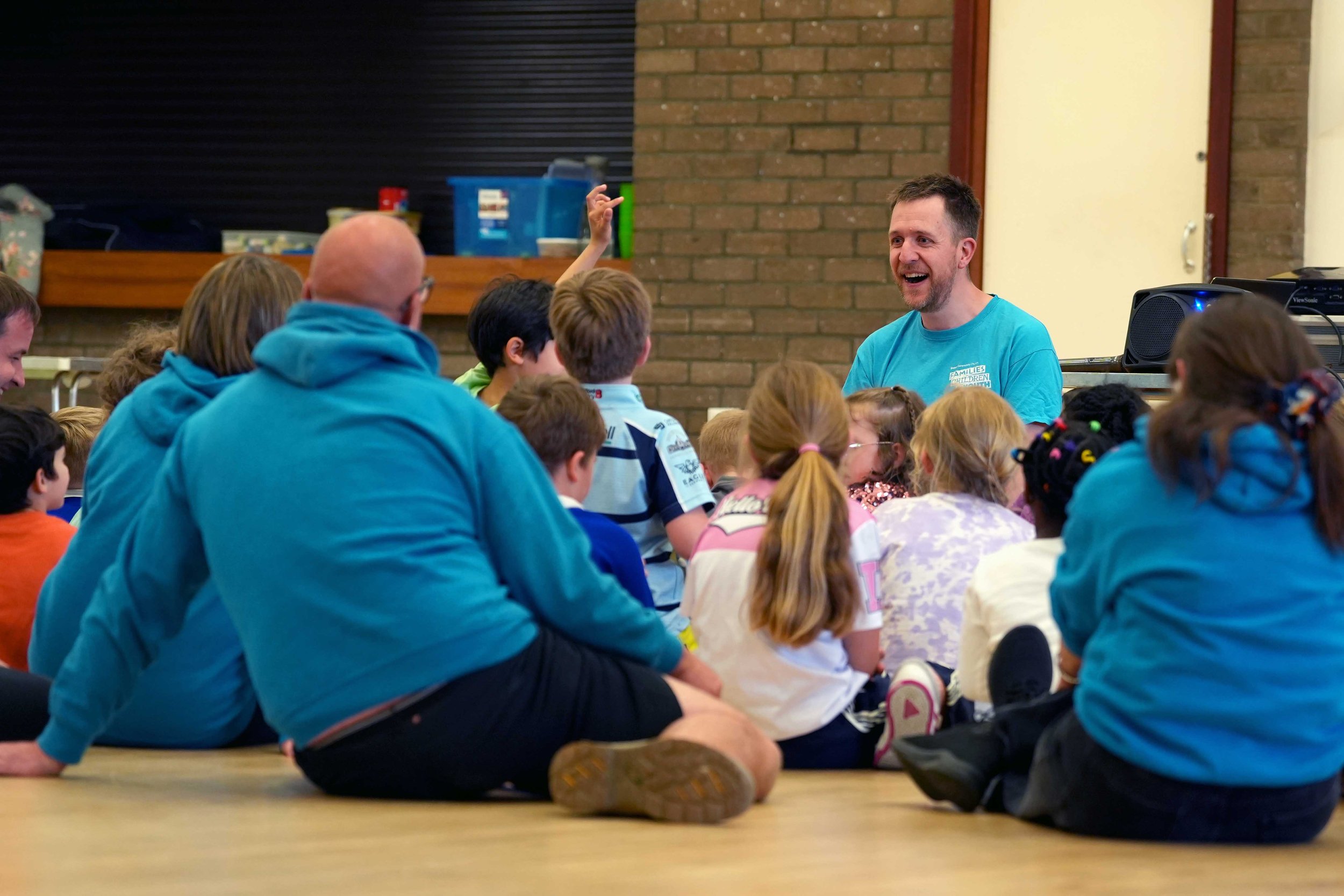 A group of children and adults sitting on the floor listening to a smiling man in a blue shirt who is speaking to them from the Bible at one of Grace's Children's clubs.
