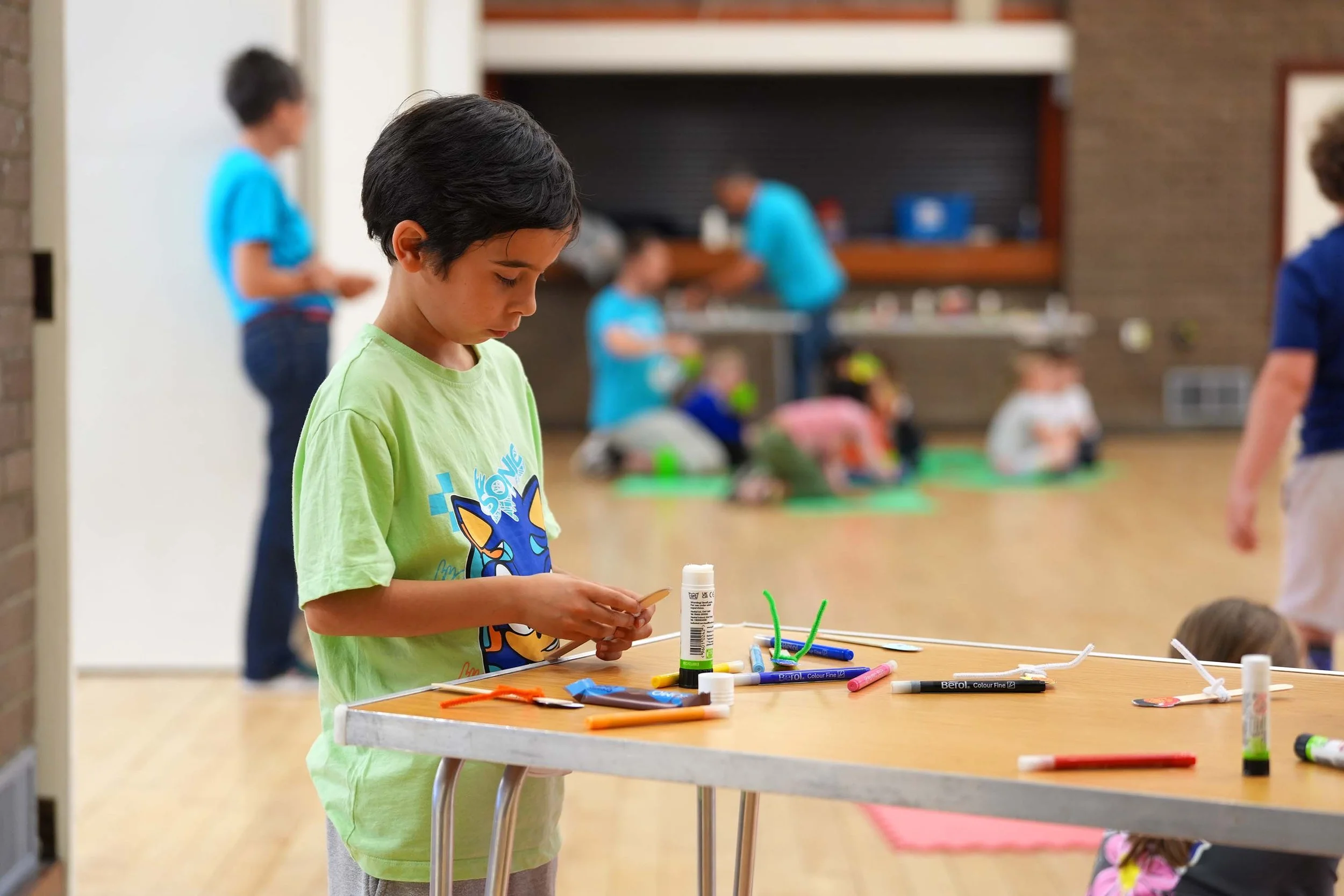 A young boy with dark hair wearing a bright green T-shirt stands at a table with colorful art supplies, including markers, glue sticks, and craft materials, in a room at one of Grace's Children's clubs